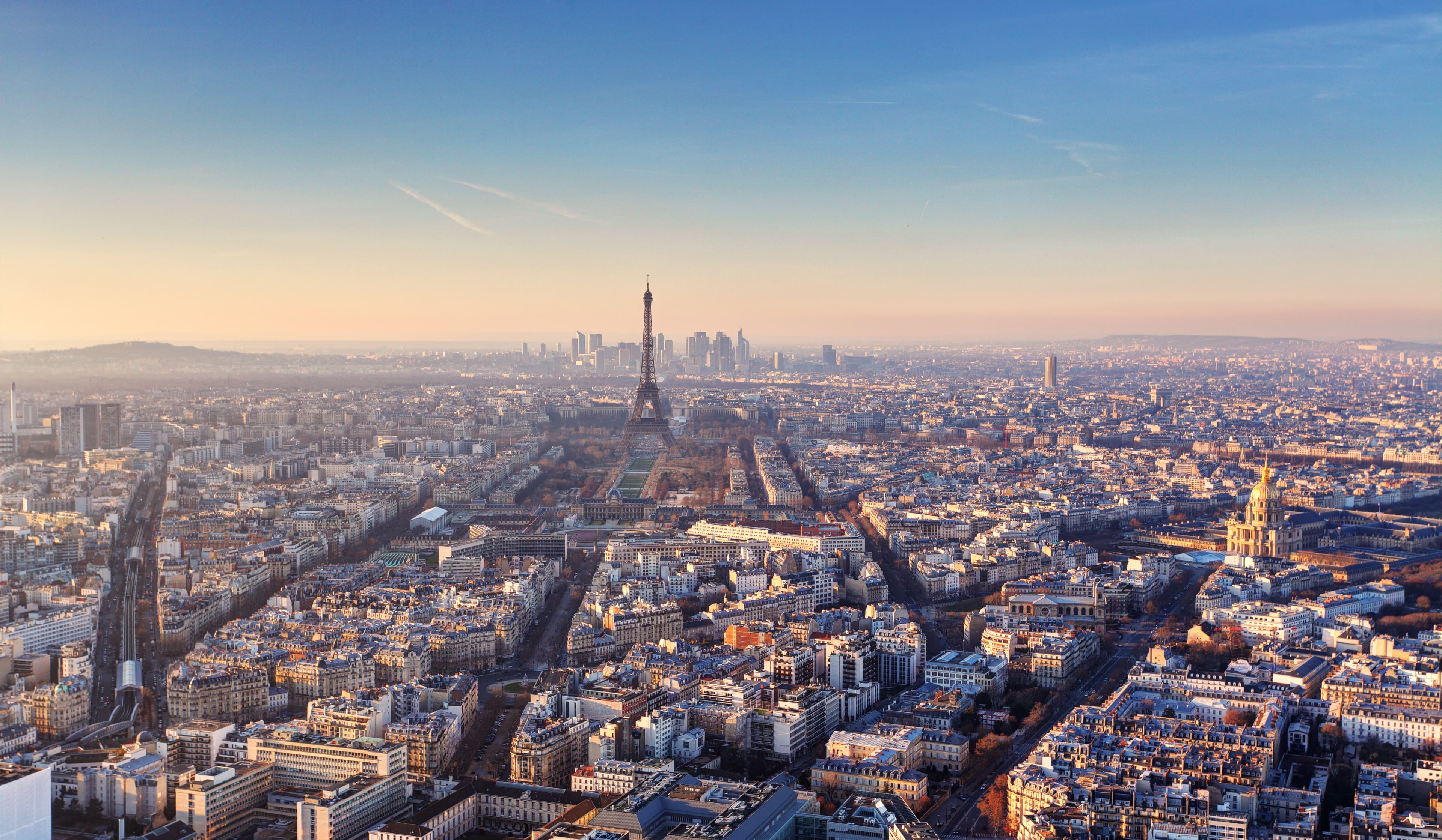 Aerial view of Paris with the Eiffel Tower in the center, surrounded by buildings and roads under a clear sky.