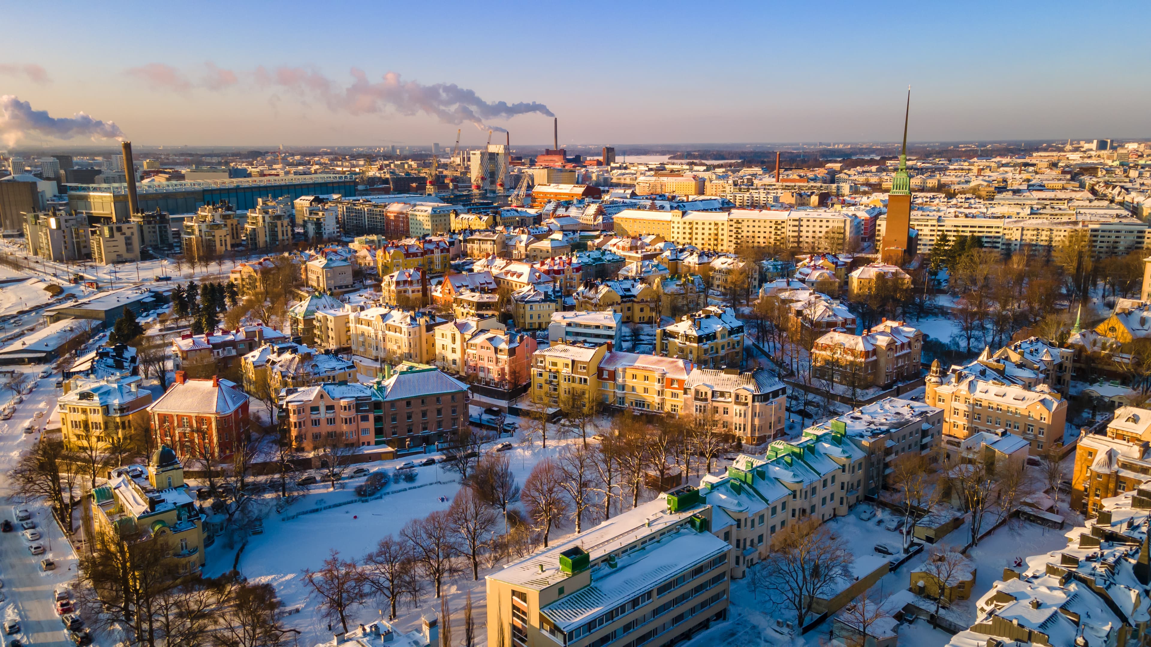 Aerial view of a cityscape with snow-covered buildings, smoke rising from industrial chimneys, and a clear blue sky in the background.