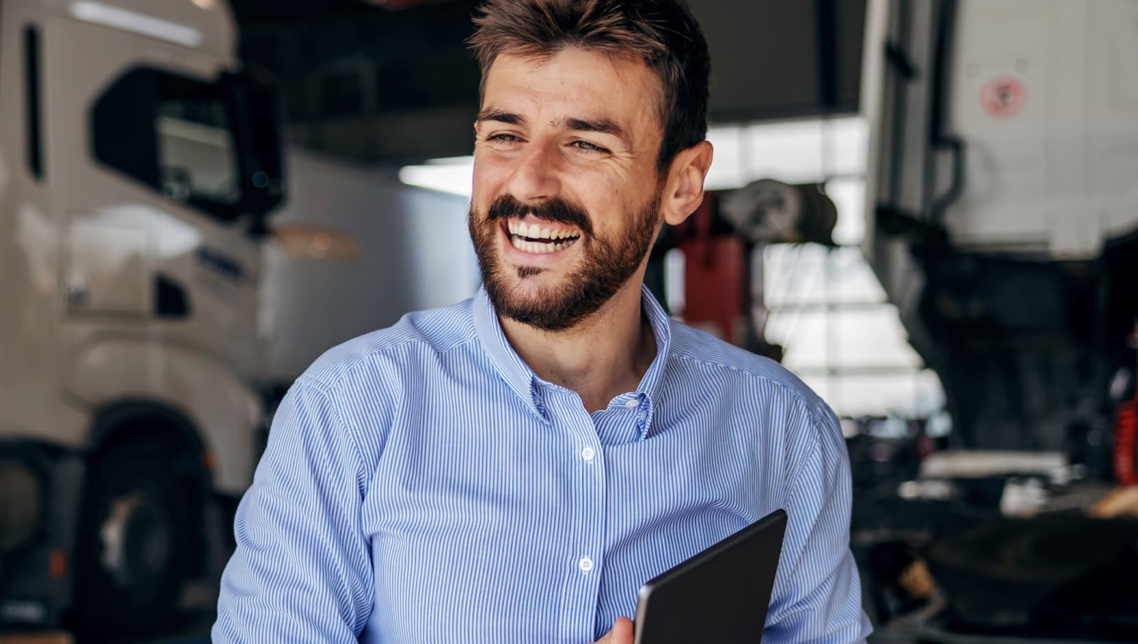 A man in a blue shirt smiles while holding a tablet in a garage with trucks in the background.