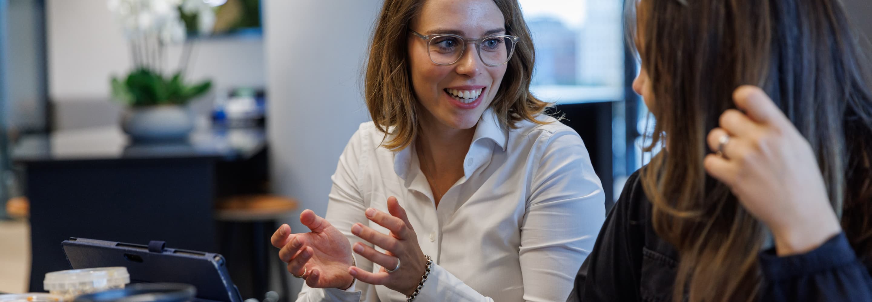 Two people are seated at a table, engaged in conversation. One person gestures with their hands while the other listens attentively. A tablet and a plant are visible in the background.