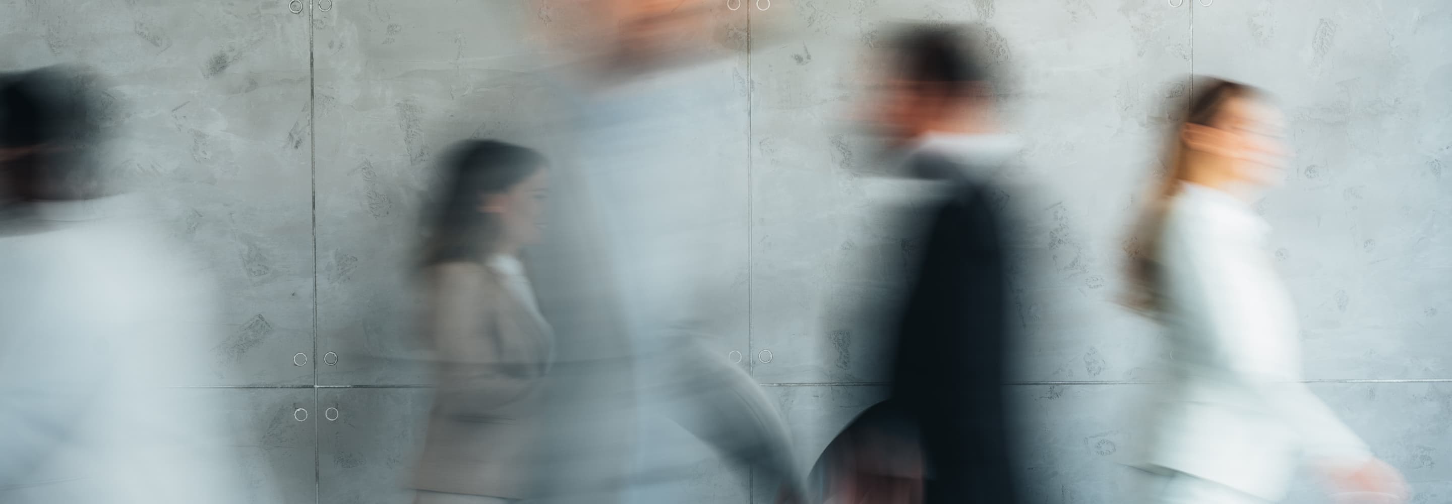 Blurred image of people walking past a gray wall, showcasing motion in an urban setting.
