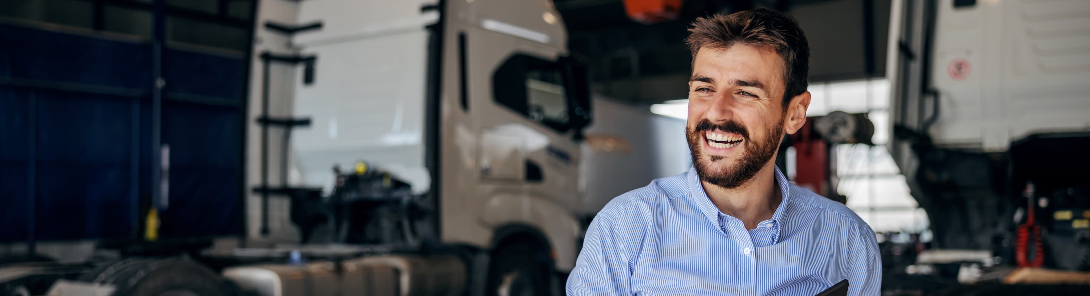 Man in a blue shirt smiling while holding a tablet in a garage with trucks in the background.