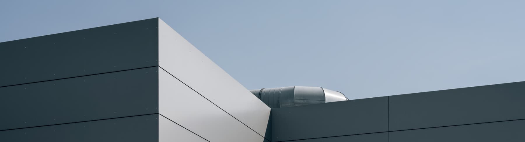 Modern building corner with smooth, gray panels against a clear blue sky; a metallic vent is partially visible above the roof.