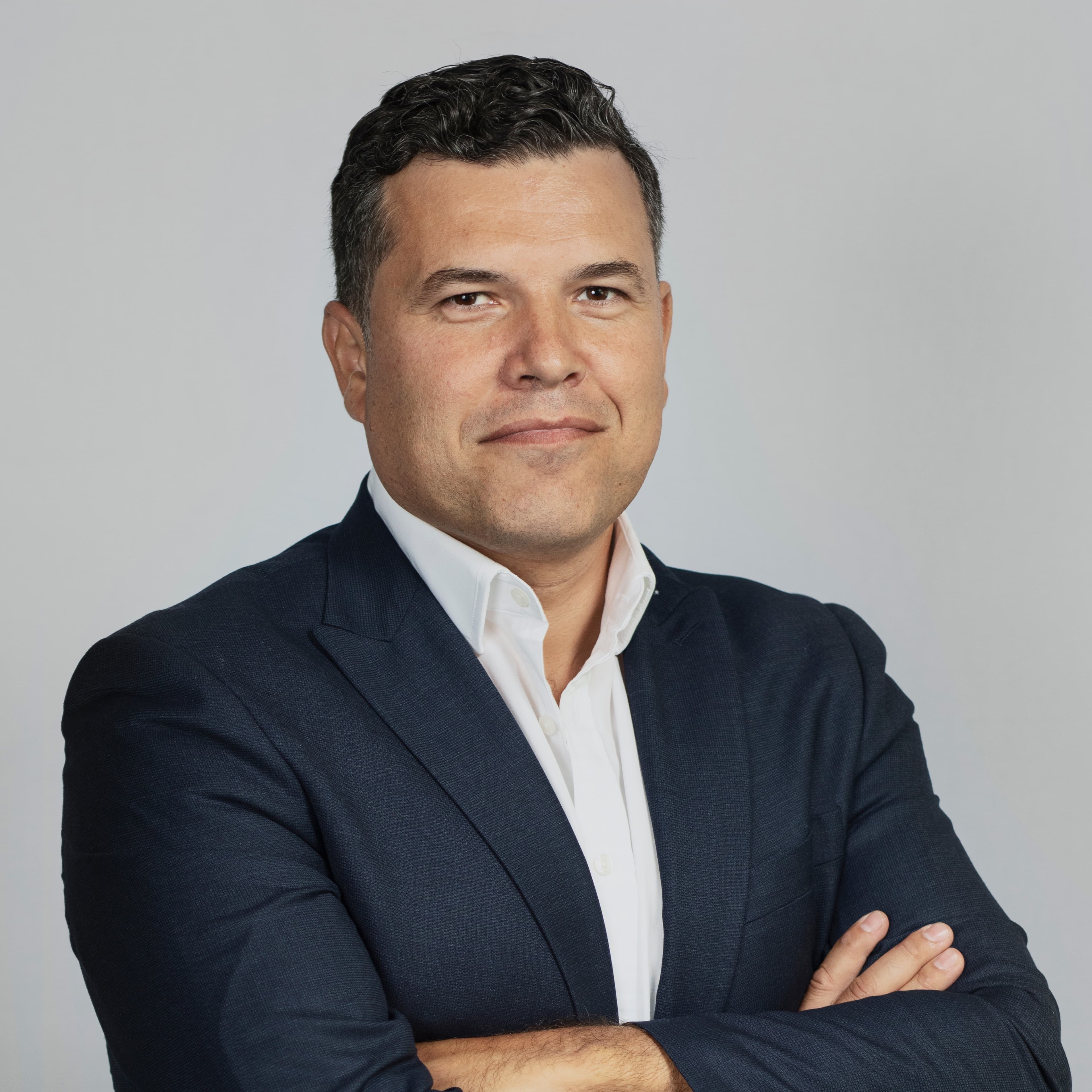 A person in a dark suit and white shirt stands with arms crossed against a plain background.
