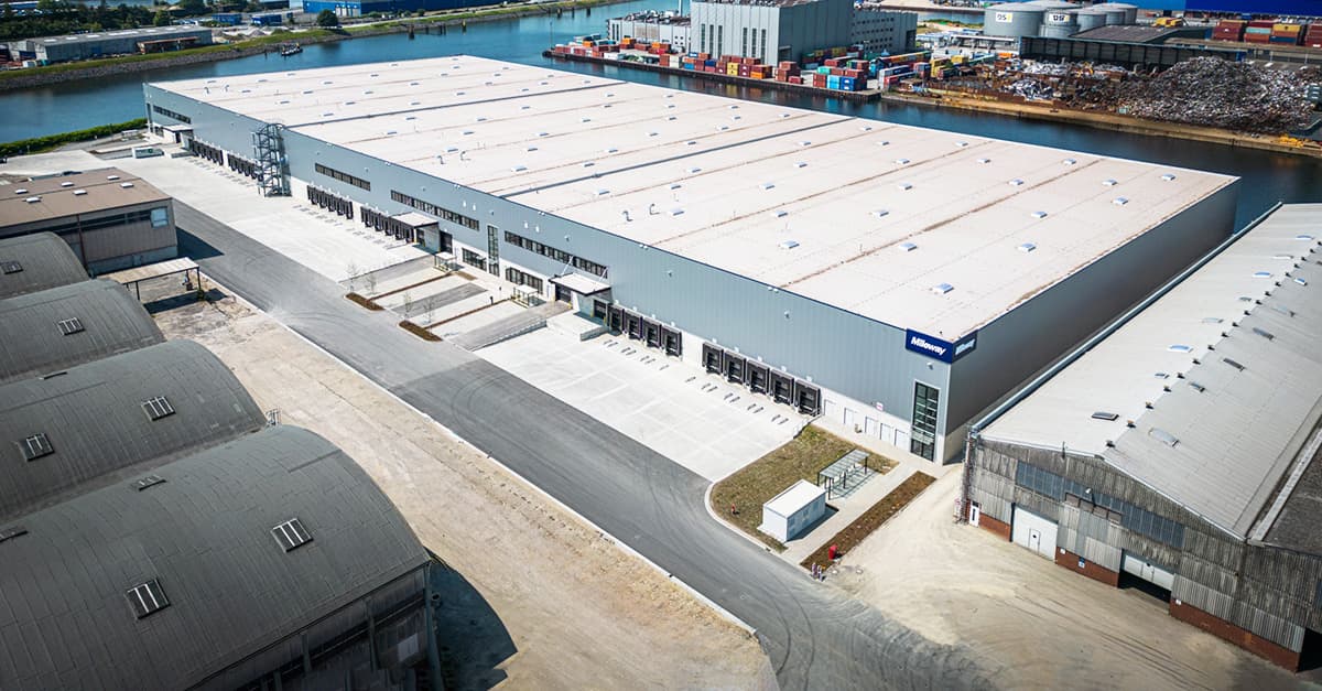 Aerial view of a large industrial warehouse with a white roof, located near a river. Surrounding structures include smaller buildings and shipping containers.