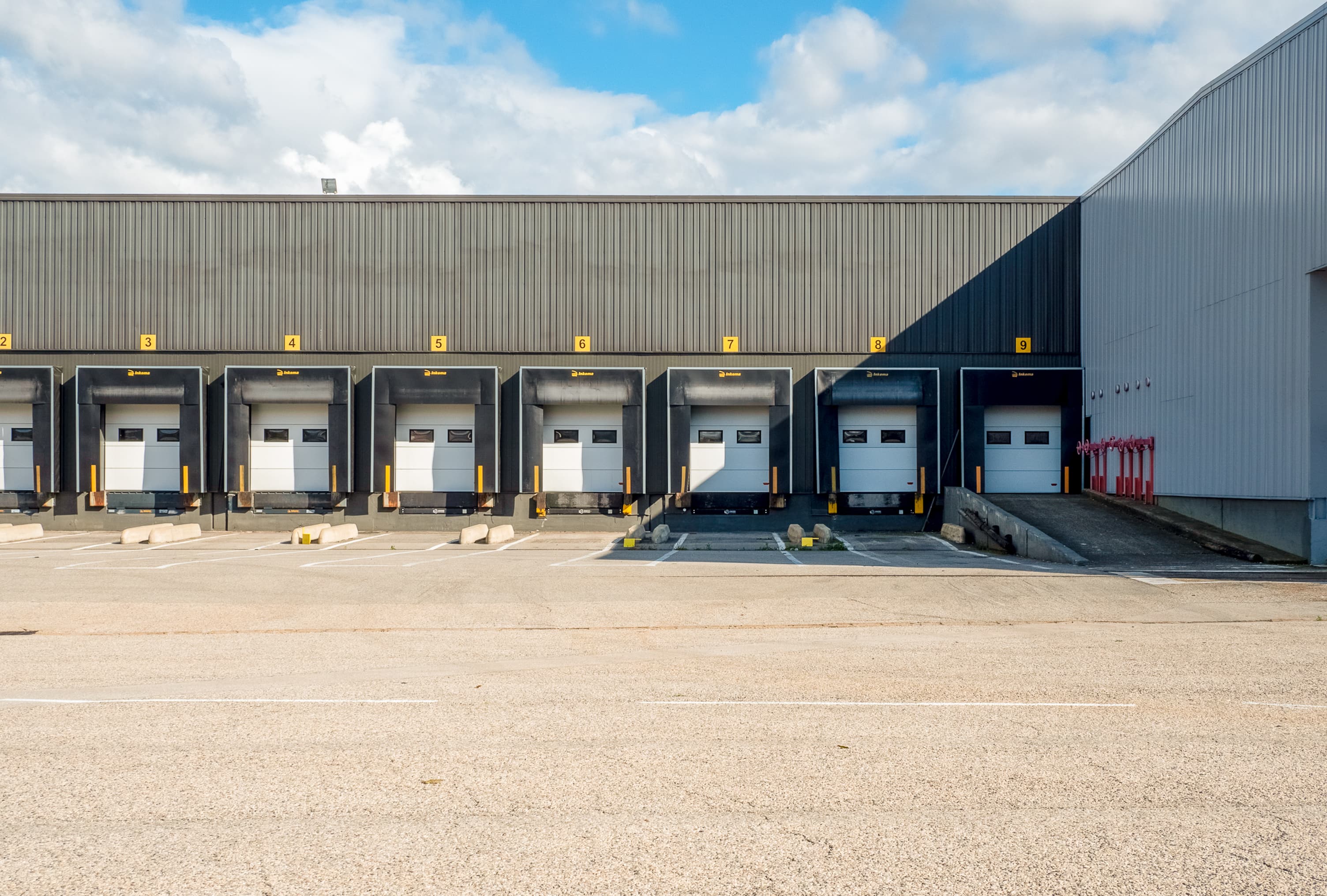 A row of closed loading dock bays at a warehouse, with a clear sky above and shadowed parking spaces in front.