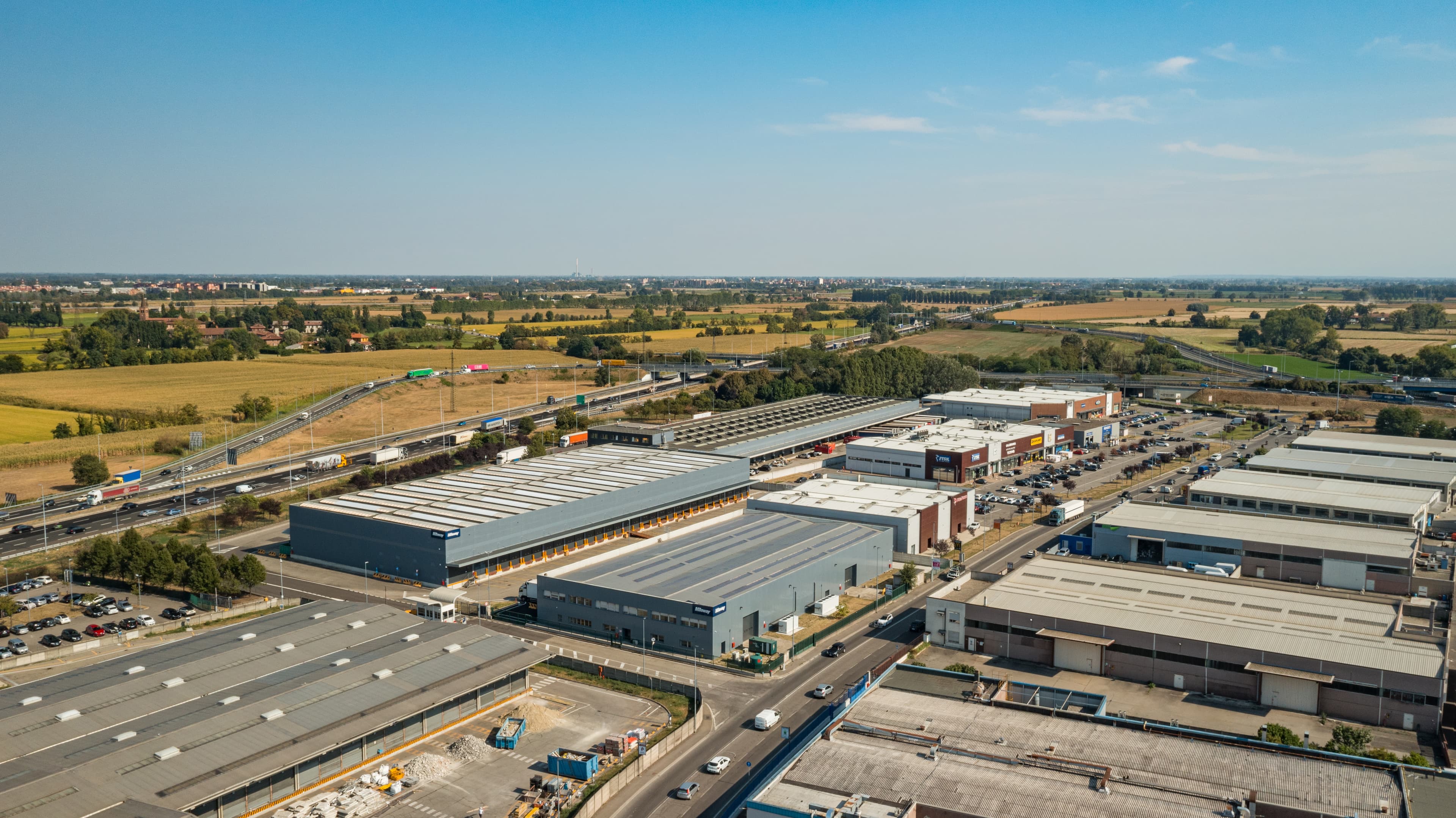 Aerial view of an industrial area with several large warehouses, roads, and vehicles. Surrounding countryside features fields and distant buildings under a clear blue sky.
