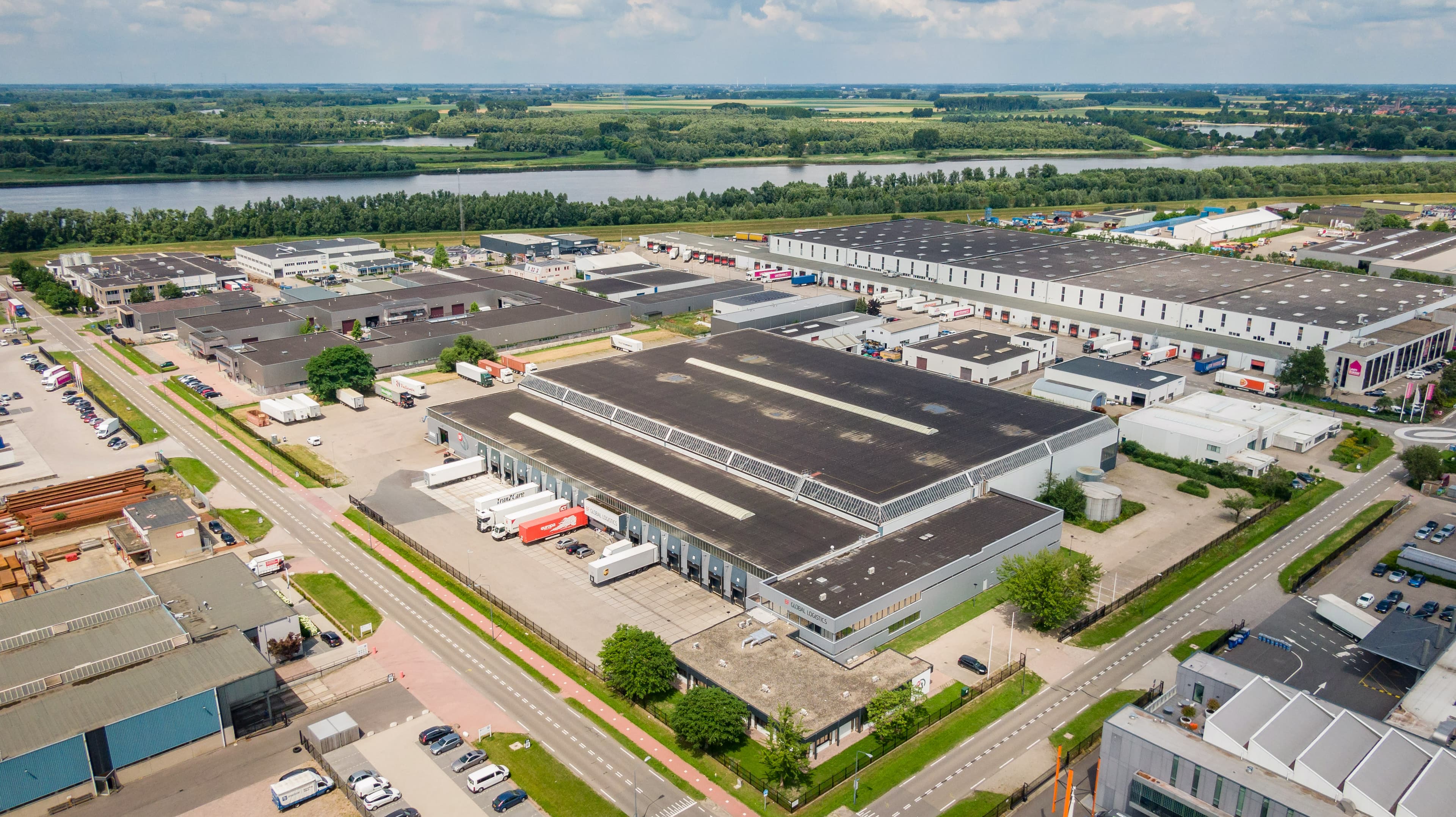 Aerial view of an industrial park with several large warehouses, trucks, and parking lots, surrounded by greenery and a river in the background.