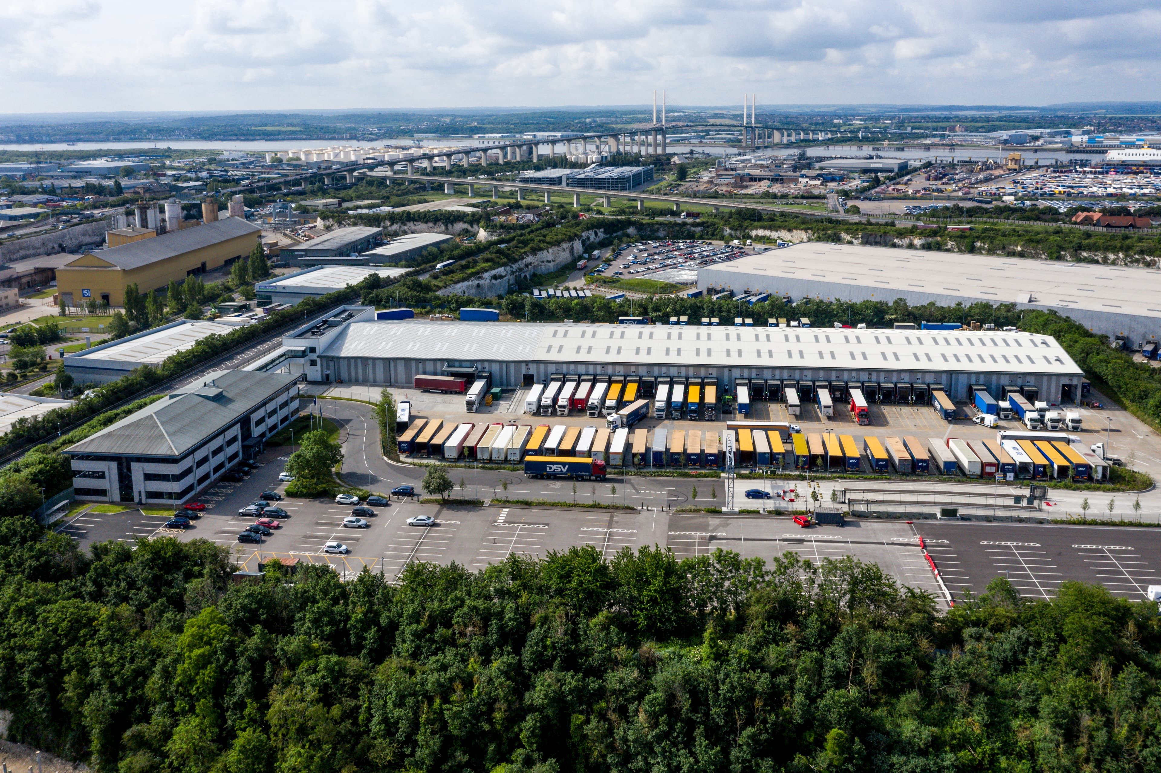 Aerial view of a large industrial warehouse with loading bays and parked trucks, surrounded by a parking lot and greenery, with a cityscape in the background under a cloudy sky.