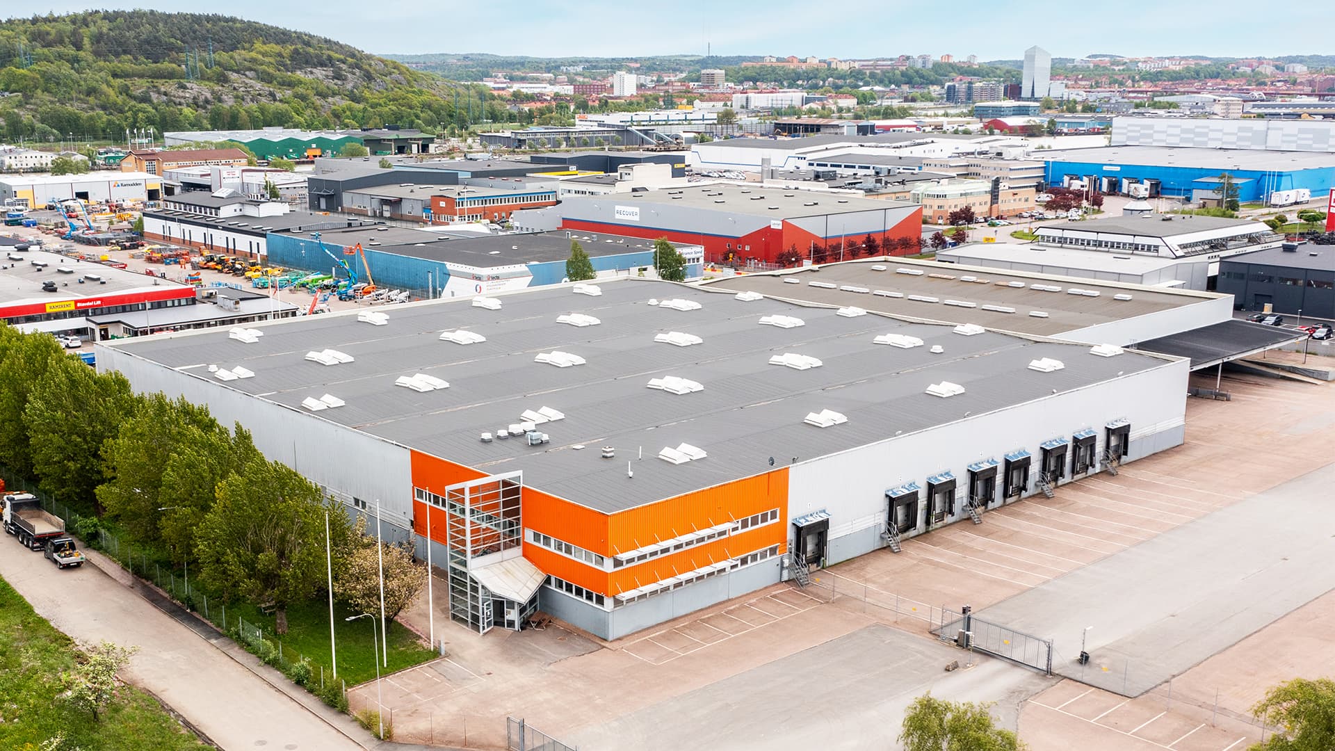 Aerial view of a large industrial warehouse with an orange and gray facade. Surrounding area features greenery, roads, and additional industrial buildings under a cloudy sky.