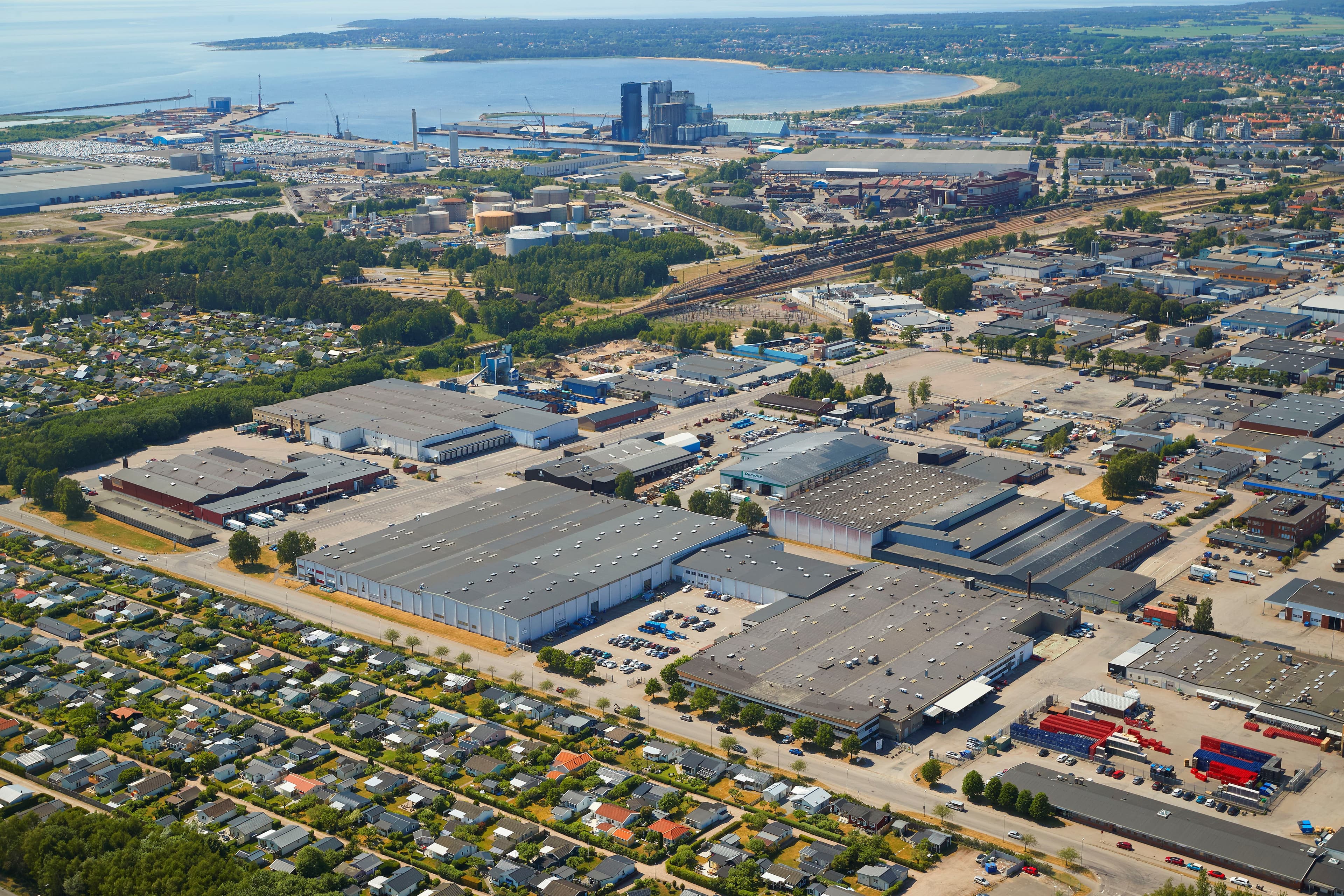 Aerial view of an industrial area near a coastline. Features warehouses, factories, a residential neighborhood, and a body of water in the background.