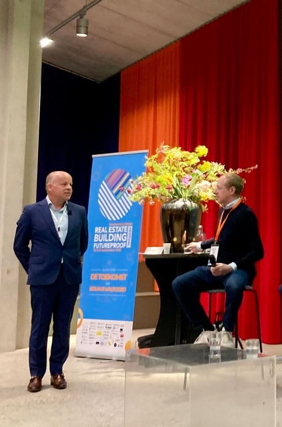 Two men are in discussion at an event. One is standing, and the other is seated by a table with a large vase of flowers. A banner in the background mentions a real estate event.