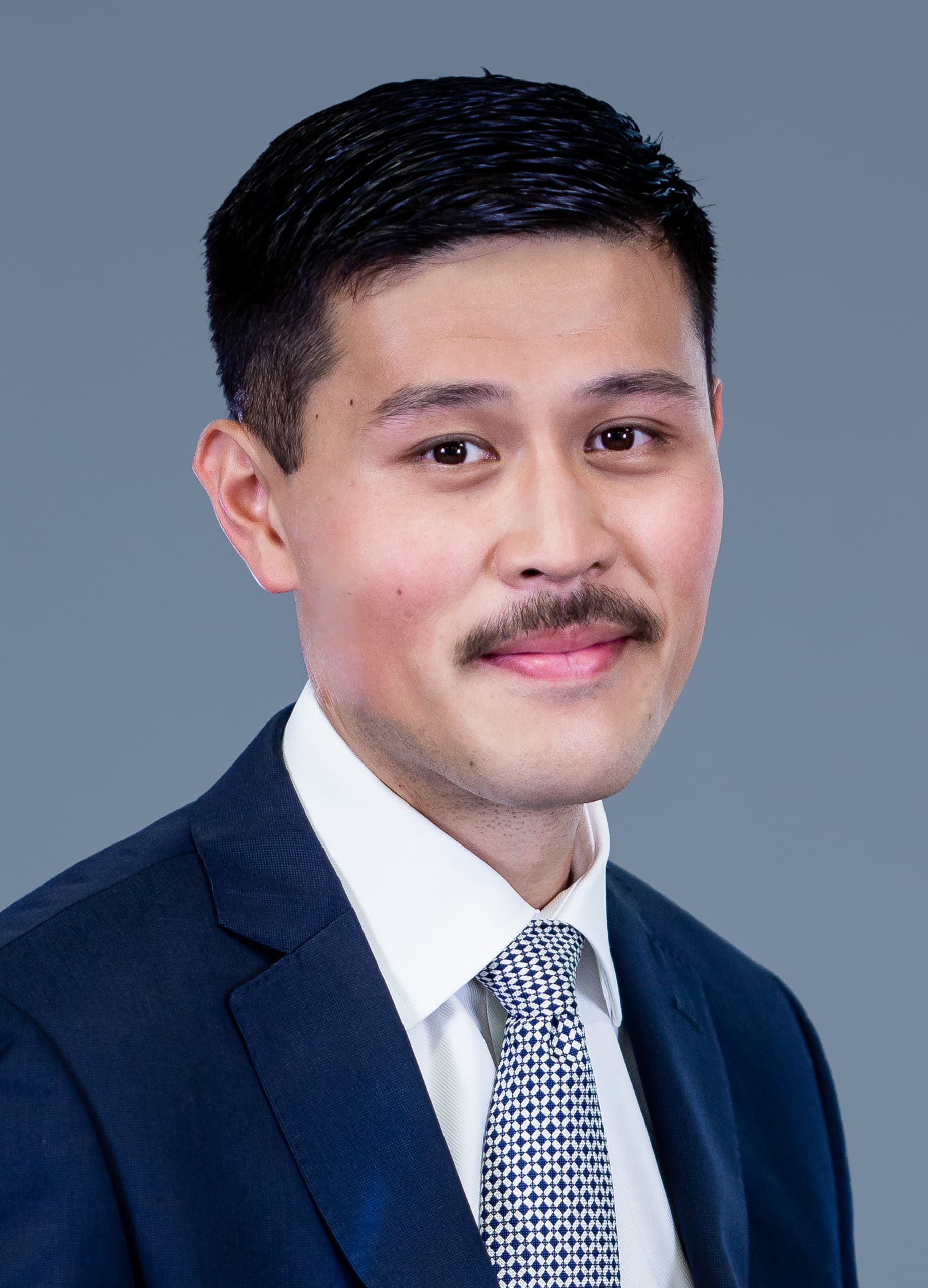 A person in a dark suit, white shirt, and patterned tie poses for a professional headshot against a plain gray background.