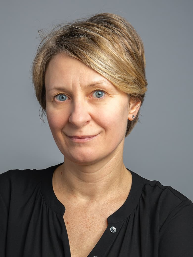 A person with short blonde hair and a slight smile is wearing a black top against a gray background.