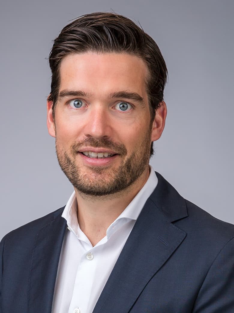 A man with dark hair and a beard, wearing a navy suit and white shirt, against a gray background.