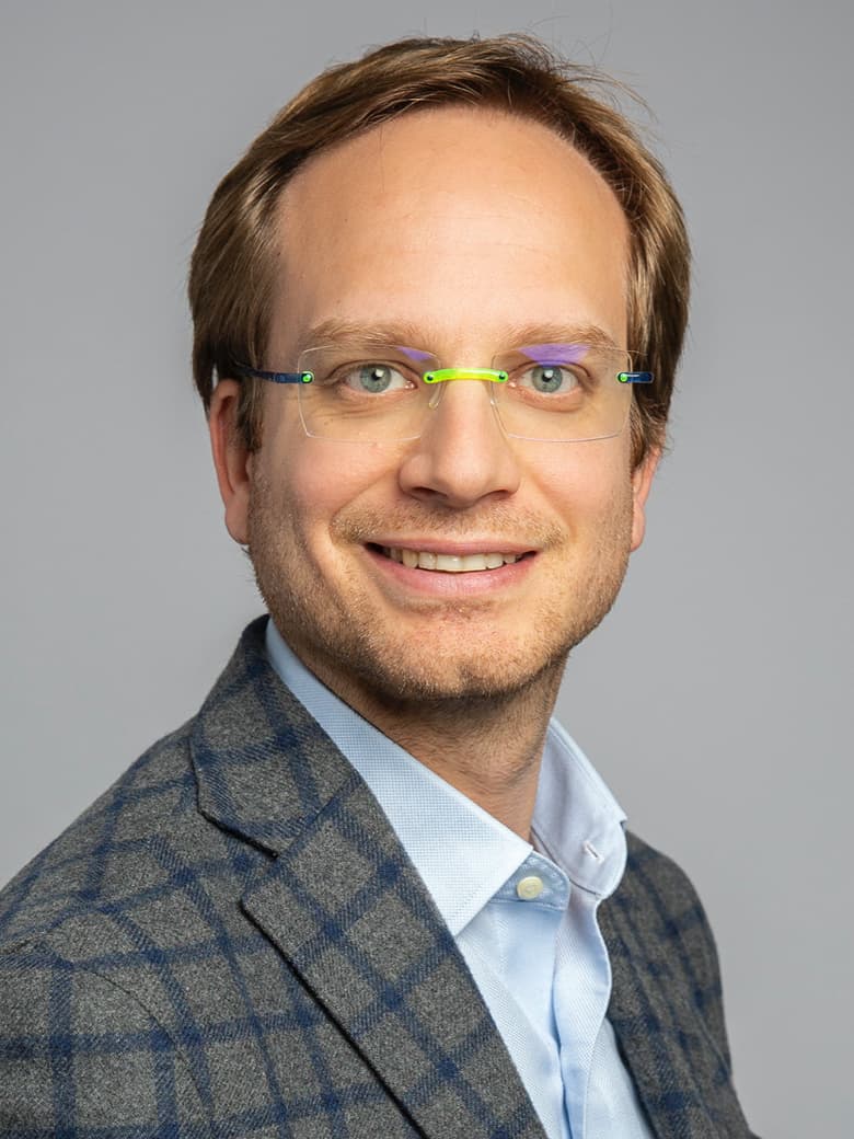 A person with light hair, wearing glasses and a checkered blazer, smiles at the camera against a neutral background.