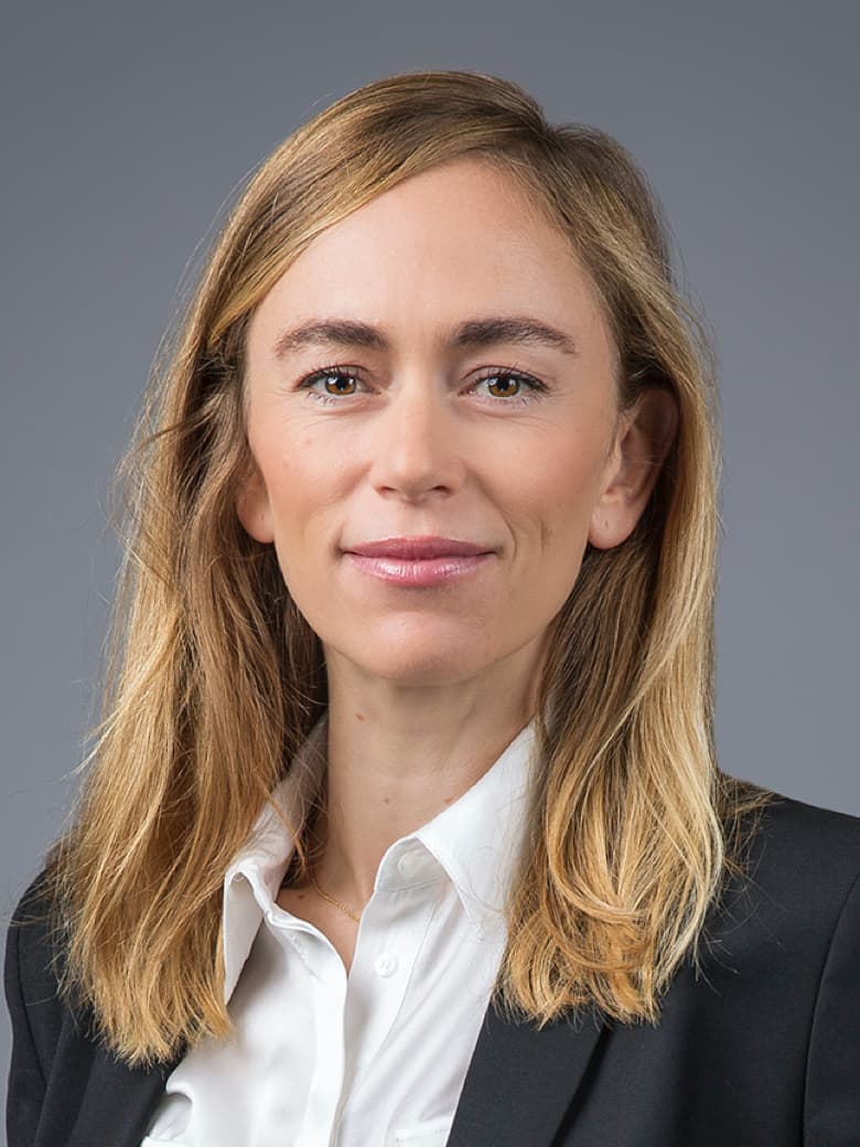 A woman with long blonde hair is wearing a black blazer and white shirt, posing against a gray background.