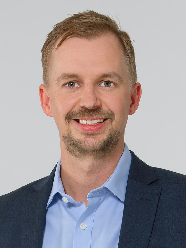 A man in a blue shirt and dark blazer smiling at the camera against a plain background.