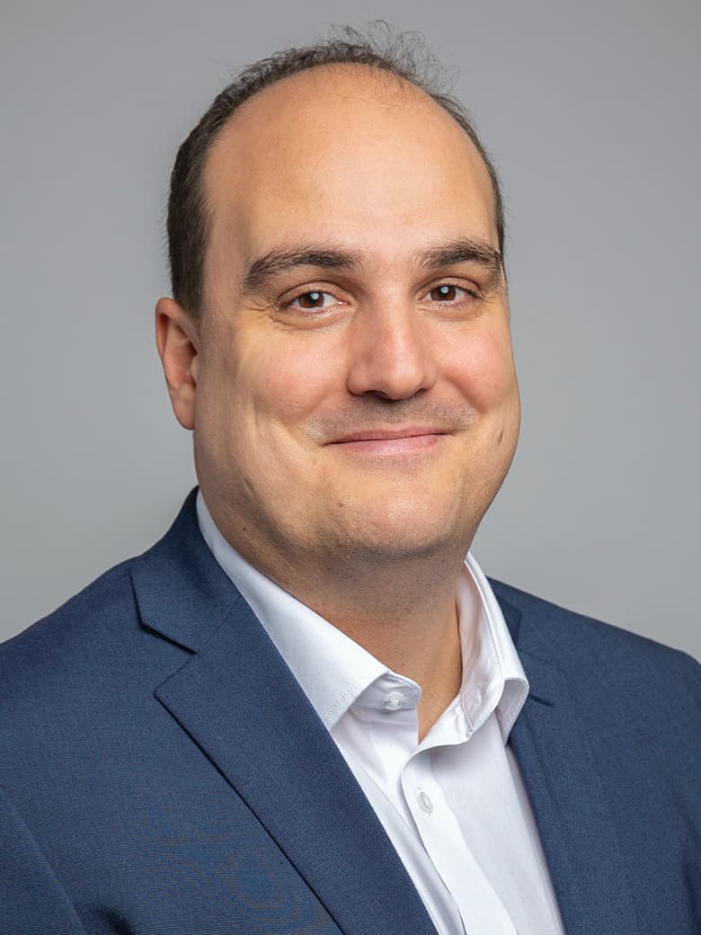 A man in a blue suit and white shirt smiles slightly against a gray background.