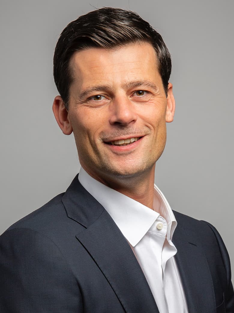 A man in a black suit and white shirt smiles at the camera against a plain gray background.