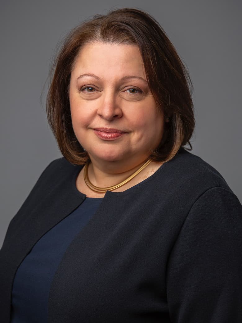 A woman with shoulder-length brown hair, wearing a dark blazer and a gold necklace, poses against a gray background.