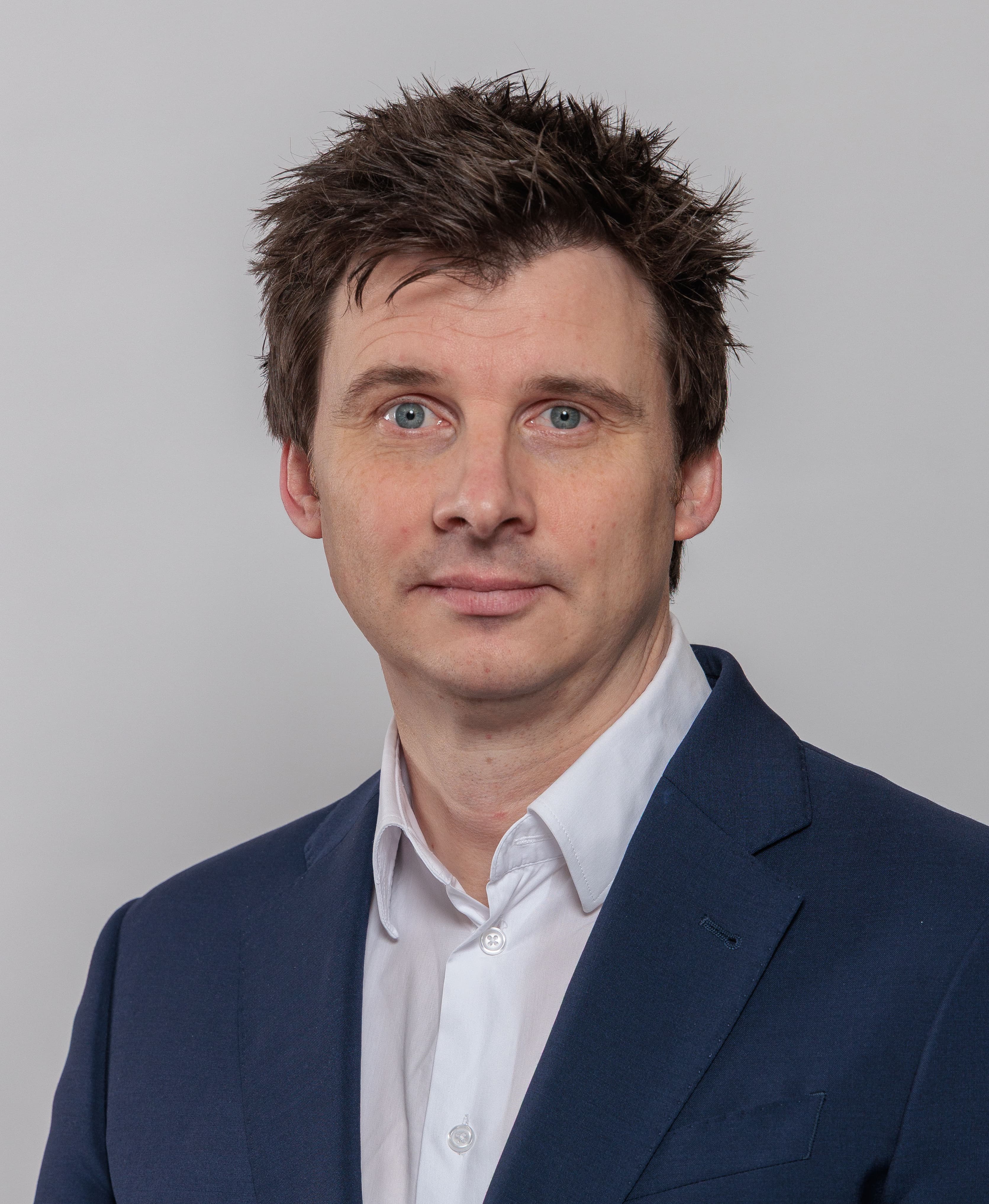 A person in a dark blue suit jacket and white shirt poses against a plain light gray background.