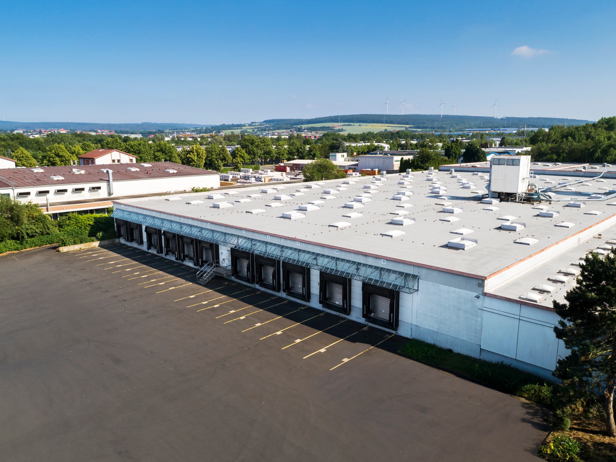 Aerial view of a large warehouse with multiple loading docks, situated in an industrial area surrounded by greenery and distant hills under a clear blue sky.