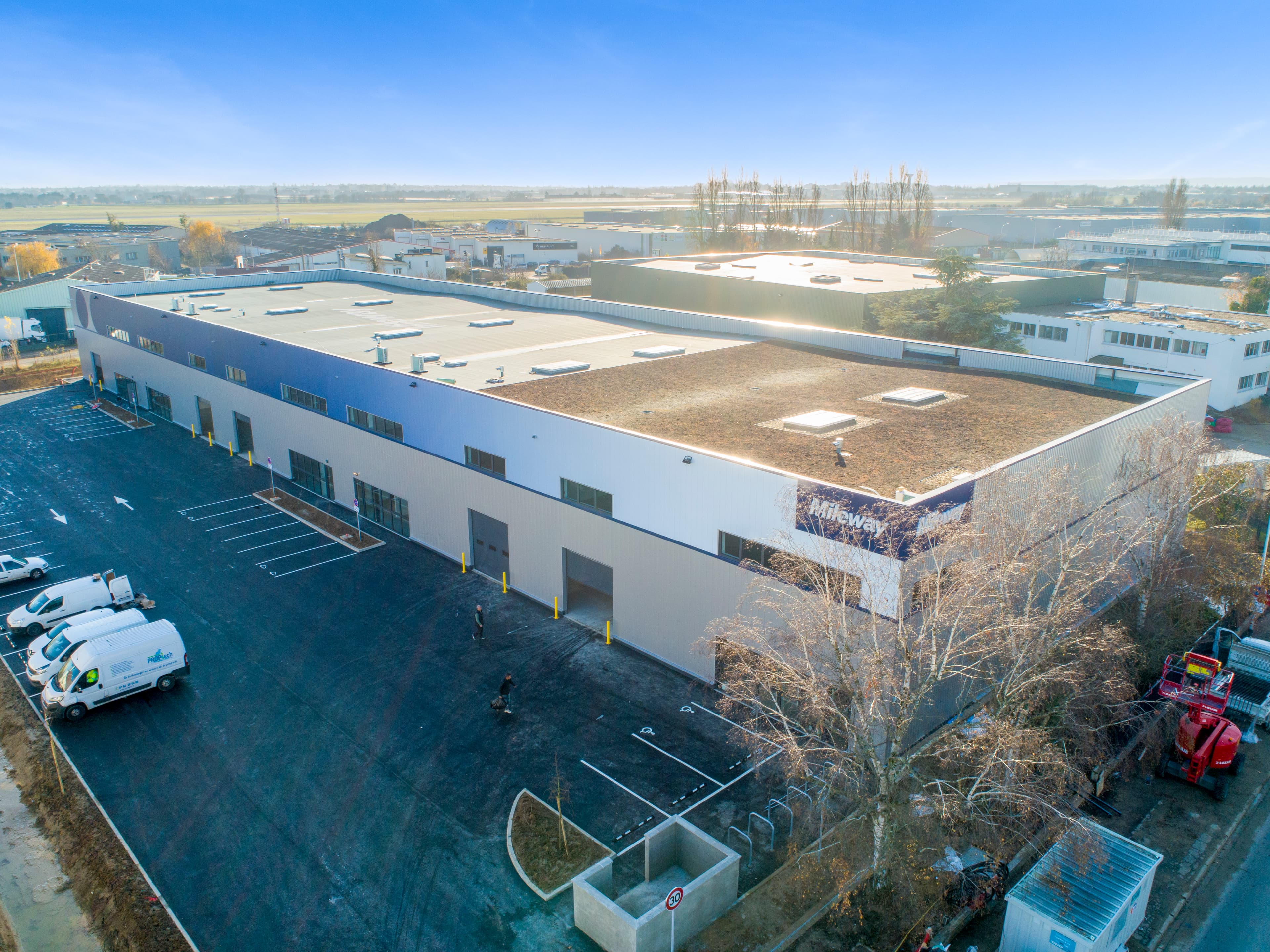 Aerial view of a large industrial warehouse with multiple loading bays, surrounded by parked vehicles and adjacent to other commercial buildings.