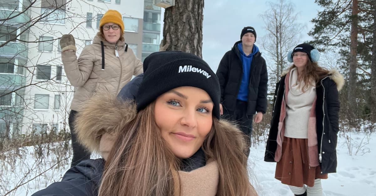 Four people in winter clothing, including hats and coats, standing outside in a snowy setting with trees and a building in the background.