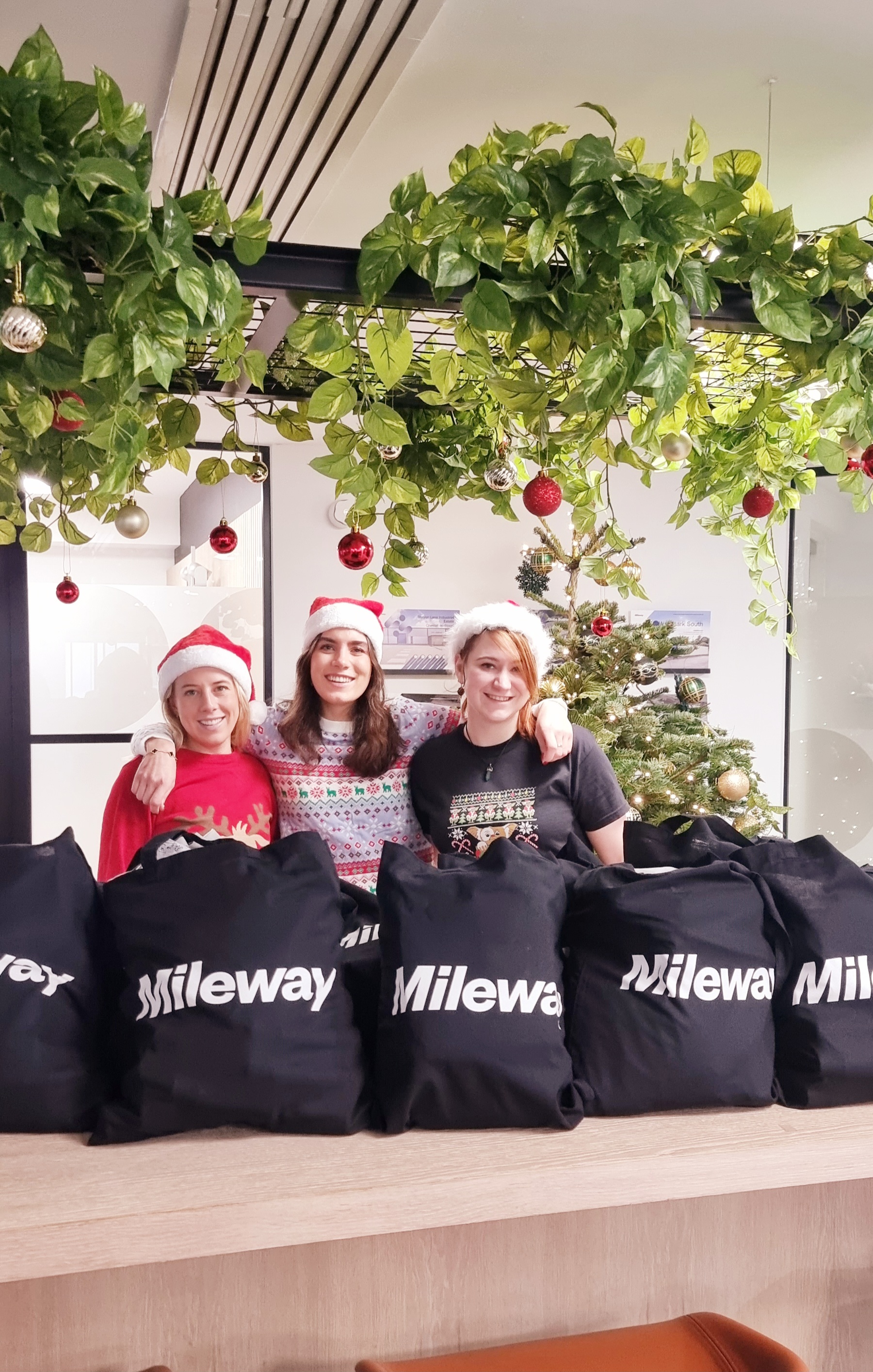 Three people wearing festive hats stand behind a table with Mileway bags. The setting is decorated with a Christmas tree and hanging green plants with ornaments.