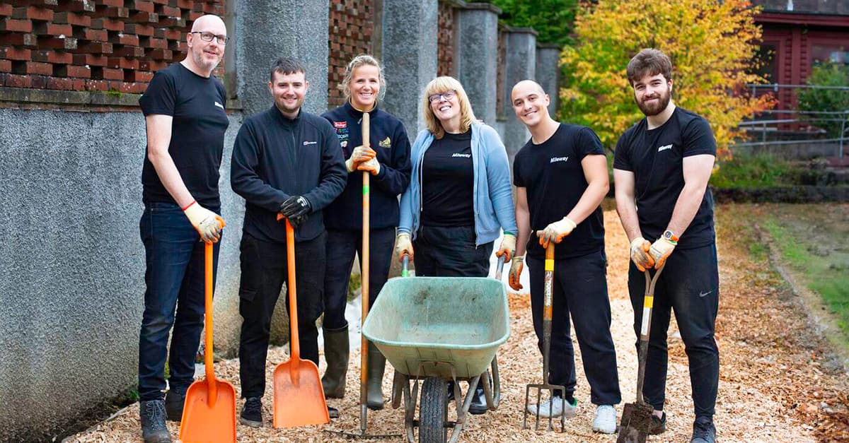 A group of six people in work attire stands with gardening tools and a wheelbarrow on a gravel path.