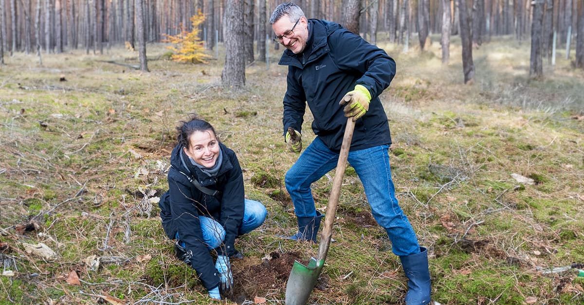 Two people, wearing jackets and gloves, planting a tree in a forest clearing. The man holds a spade, while the woman kneels, smiling.