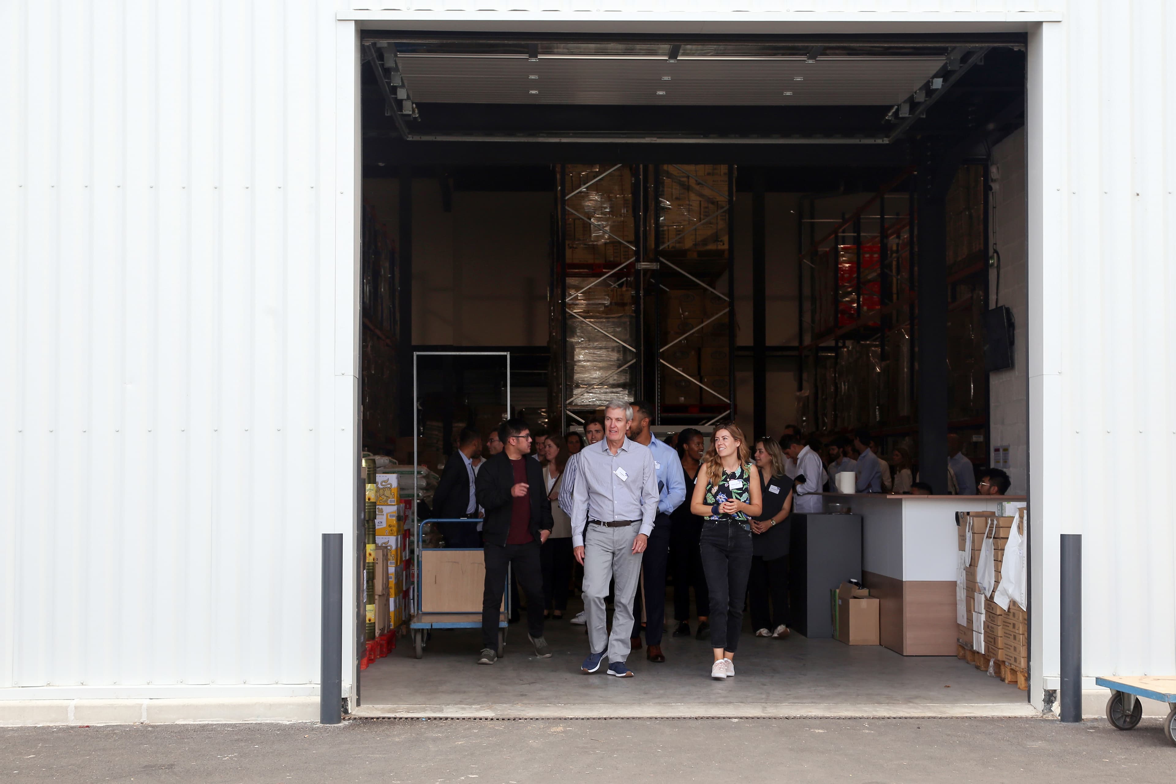 A group of people walks out of a warehouse with shelves of boxes inside. A man and a woman lead the group.