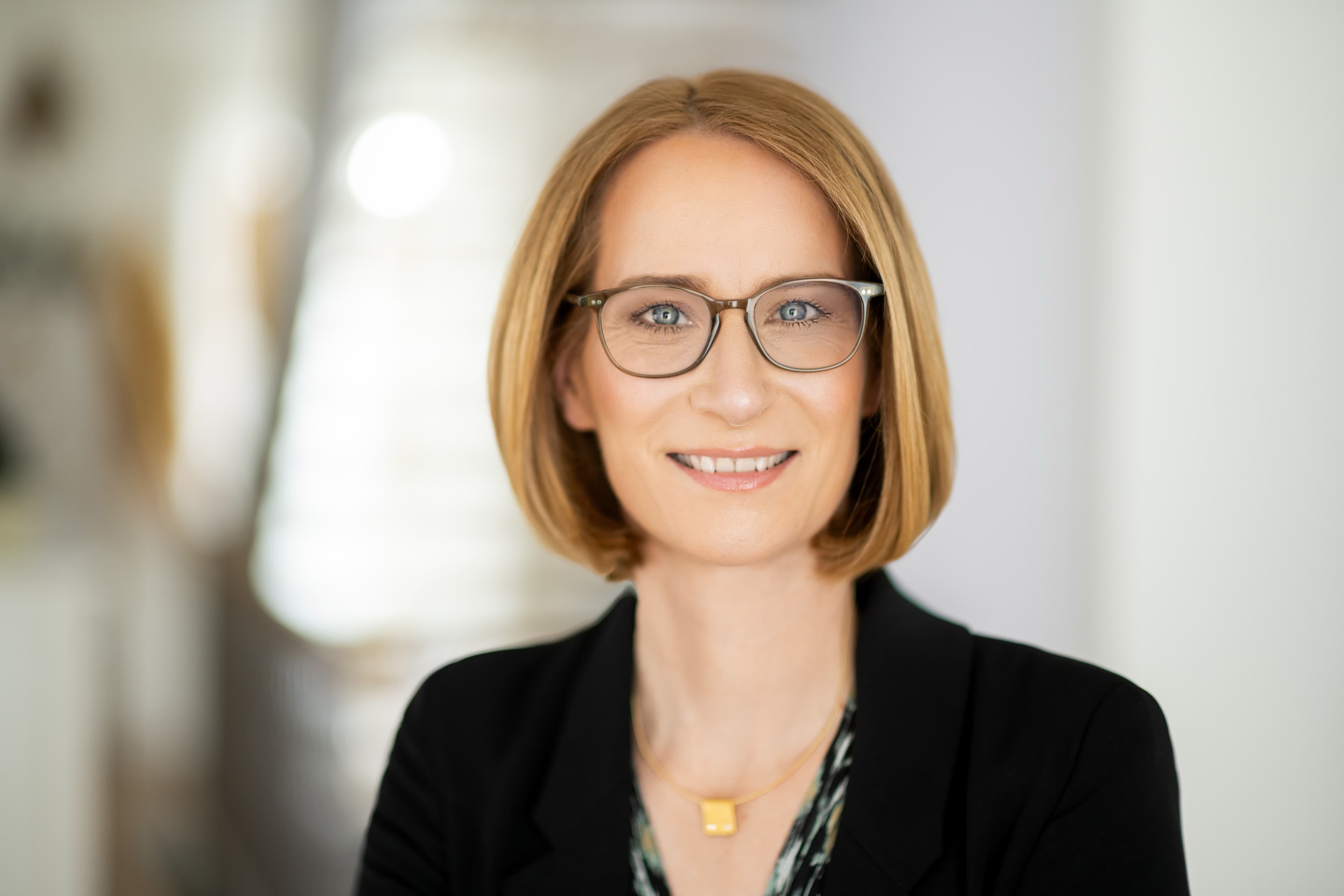 A person with short blonde hair and glasses smiles, wearing a black blazer and a necklace. The background is softly blurred.