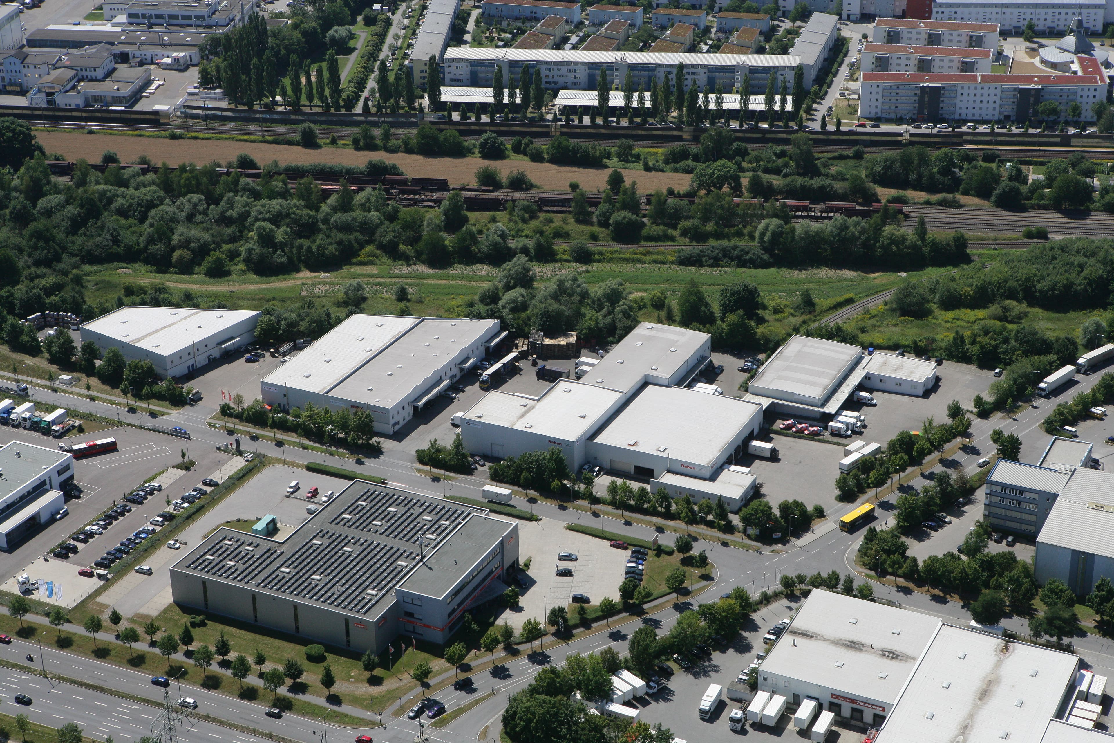 Aerial view of an industrial area with multiple white warehouse buildings, surrounded by roads, trees, and adjacent residential neighborhoods.
