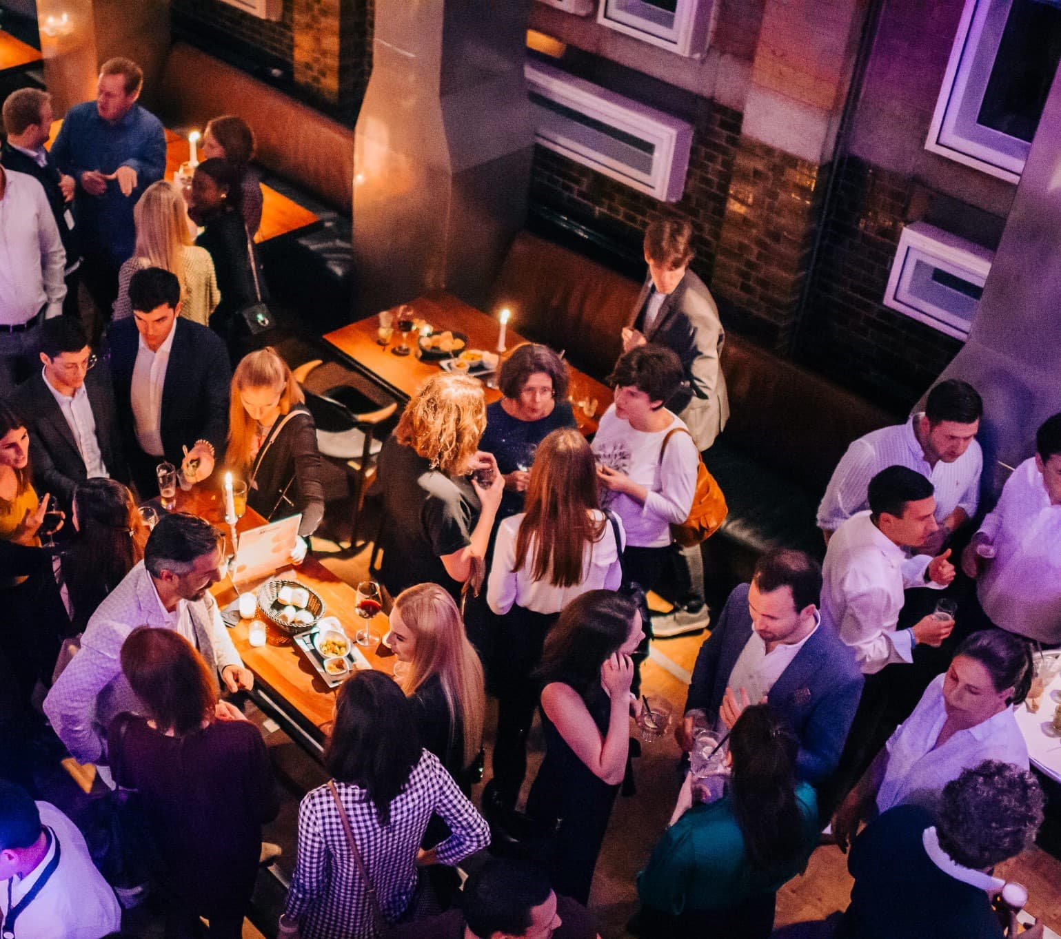 A crowded indoor gathering with people socializing around tables, some holding drinks. The setting has warm lighting and wooden furniture.
