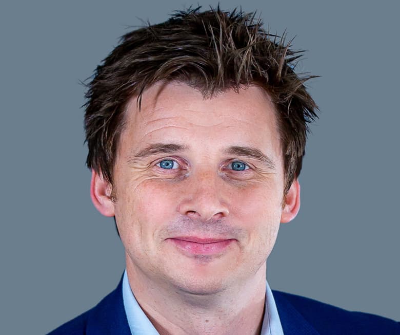 A person with short hair wearing a blue blazer against a gray background.