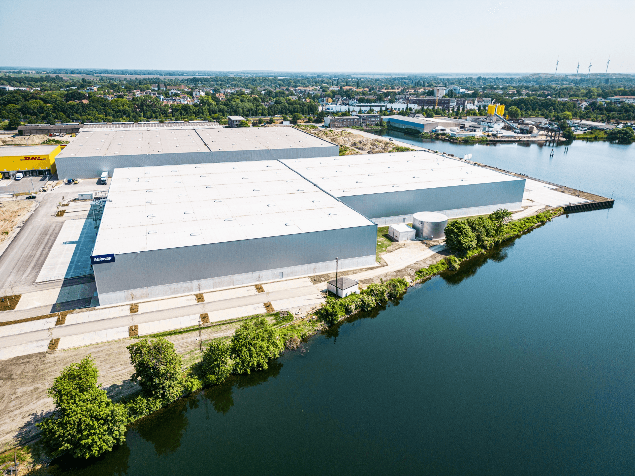 Aerial view of a large industrial warehouse complex by a river, surrounded by greenery and adjacent to other industrial facilities and buildings.