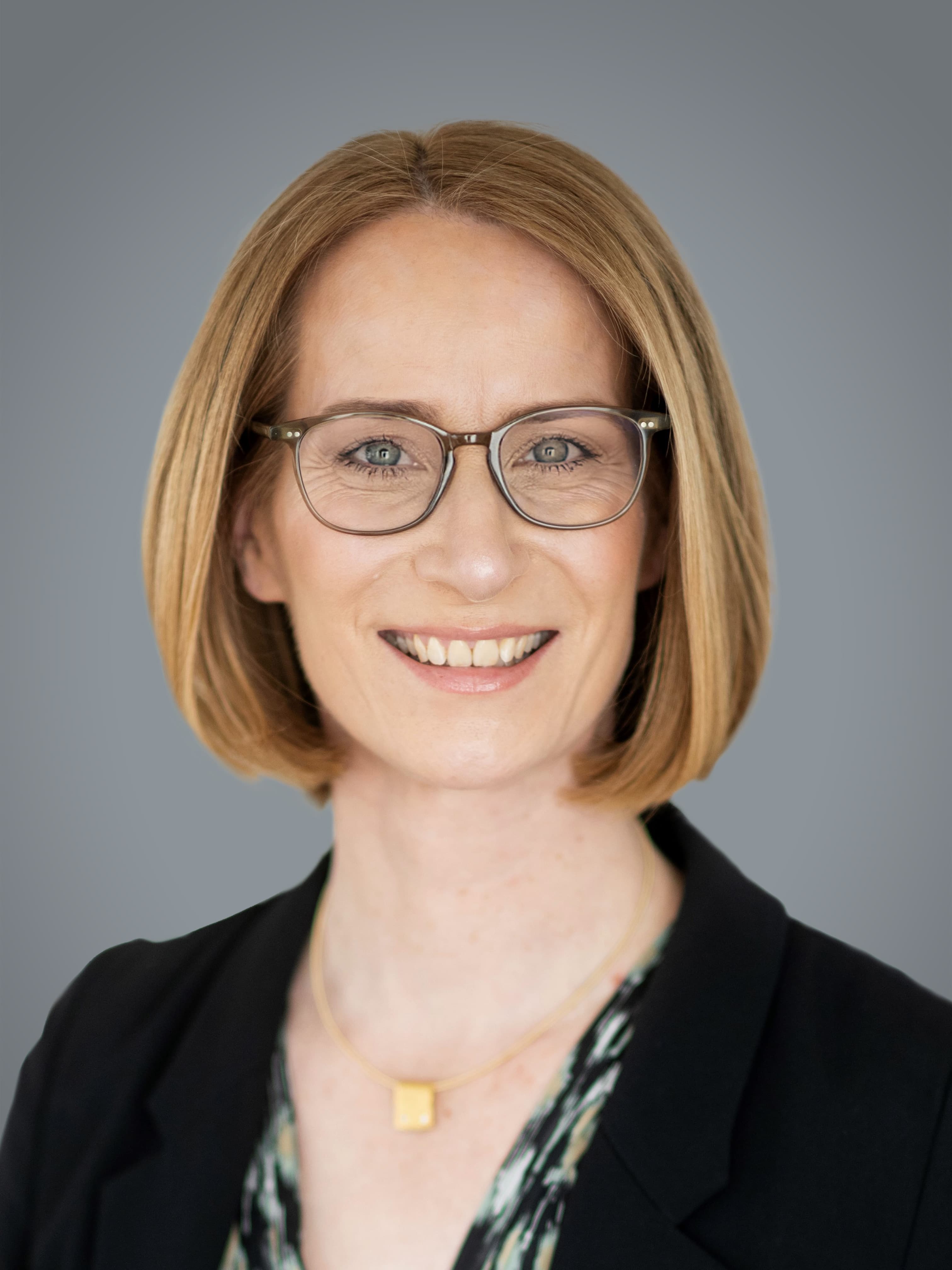 A person wearing glasses, a black blazer, and a patterned blouse smiles at the camera against a plain gray background.