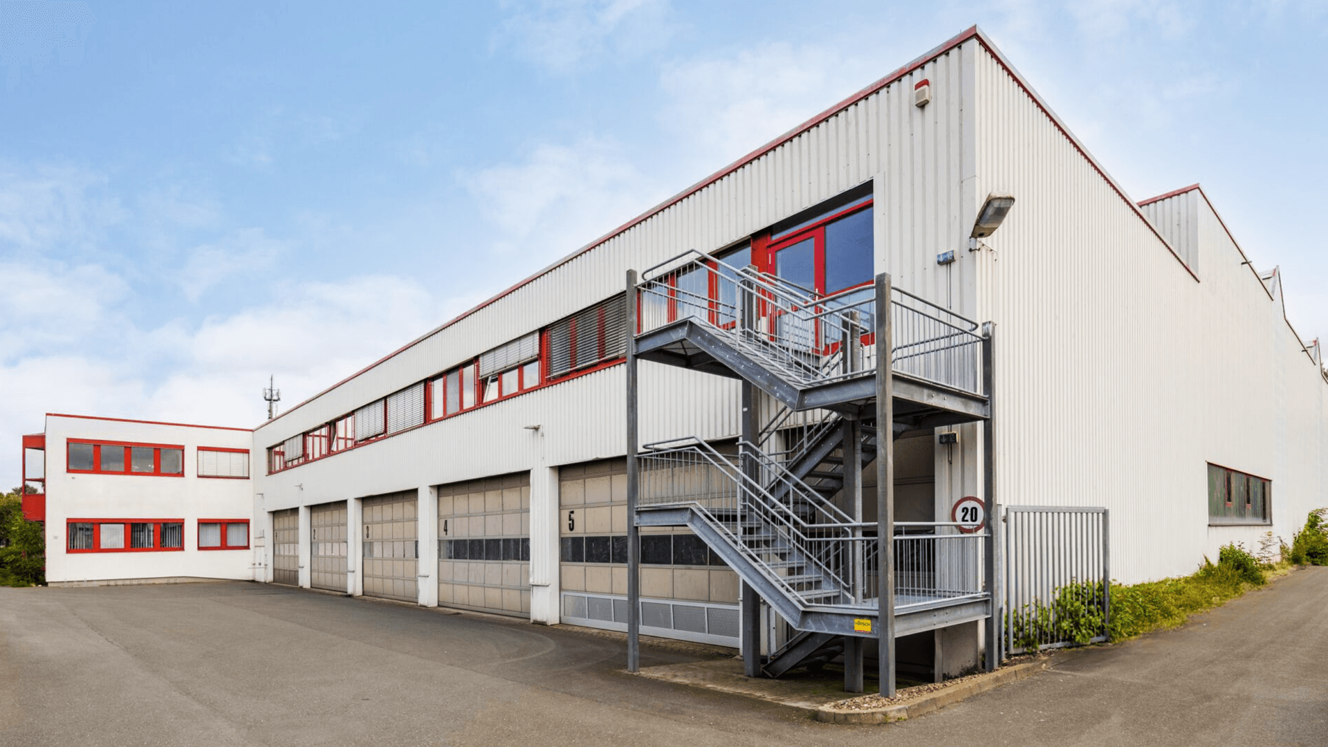 White industrial building with red trim features multiple garage doors and a metal external staircase. The sky is clear and blue.