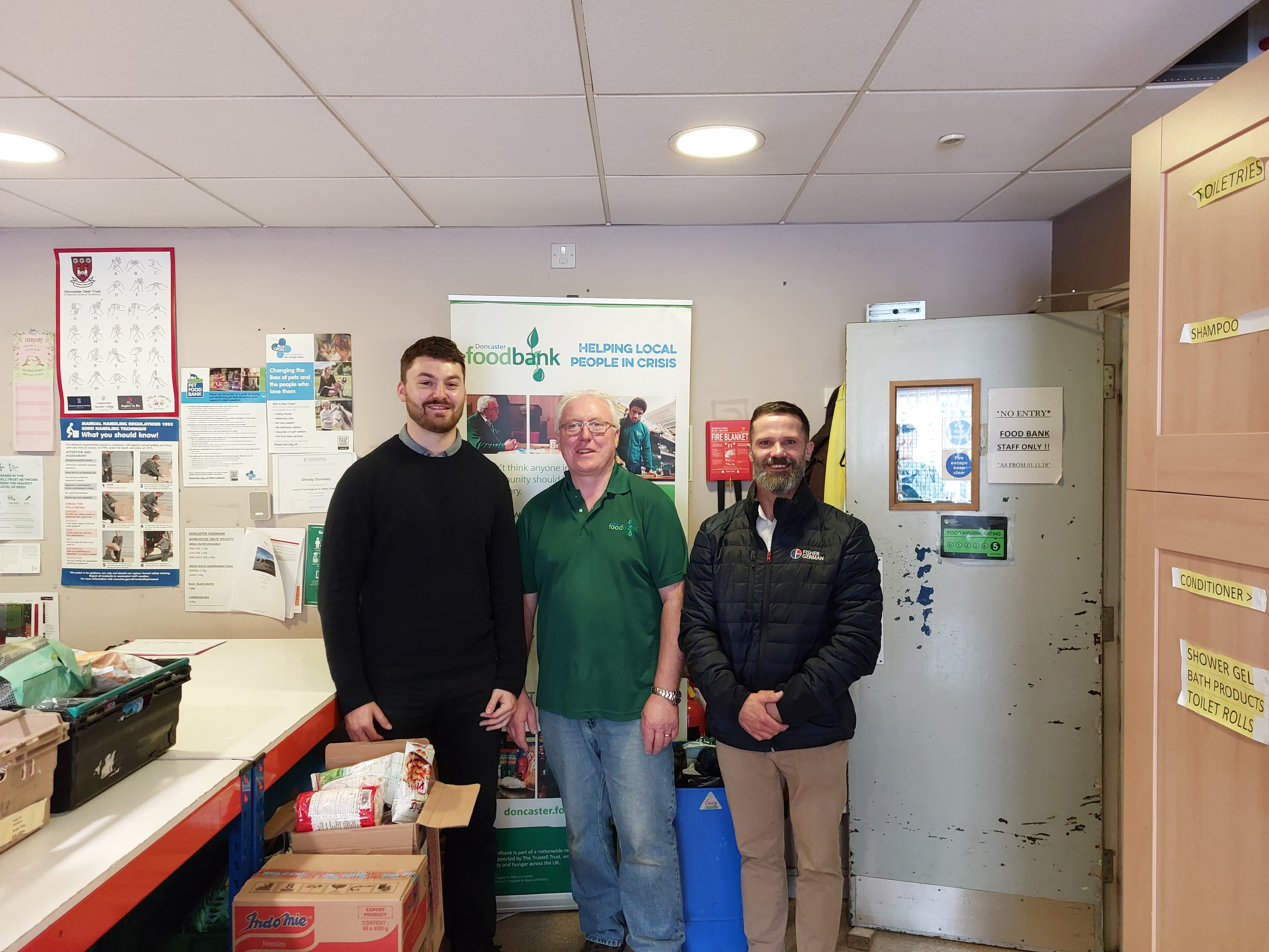 Three men stand in a food bank with posters and boxes. They are smiling and posing for a group photo.
