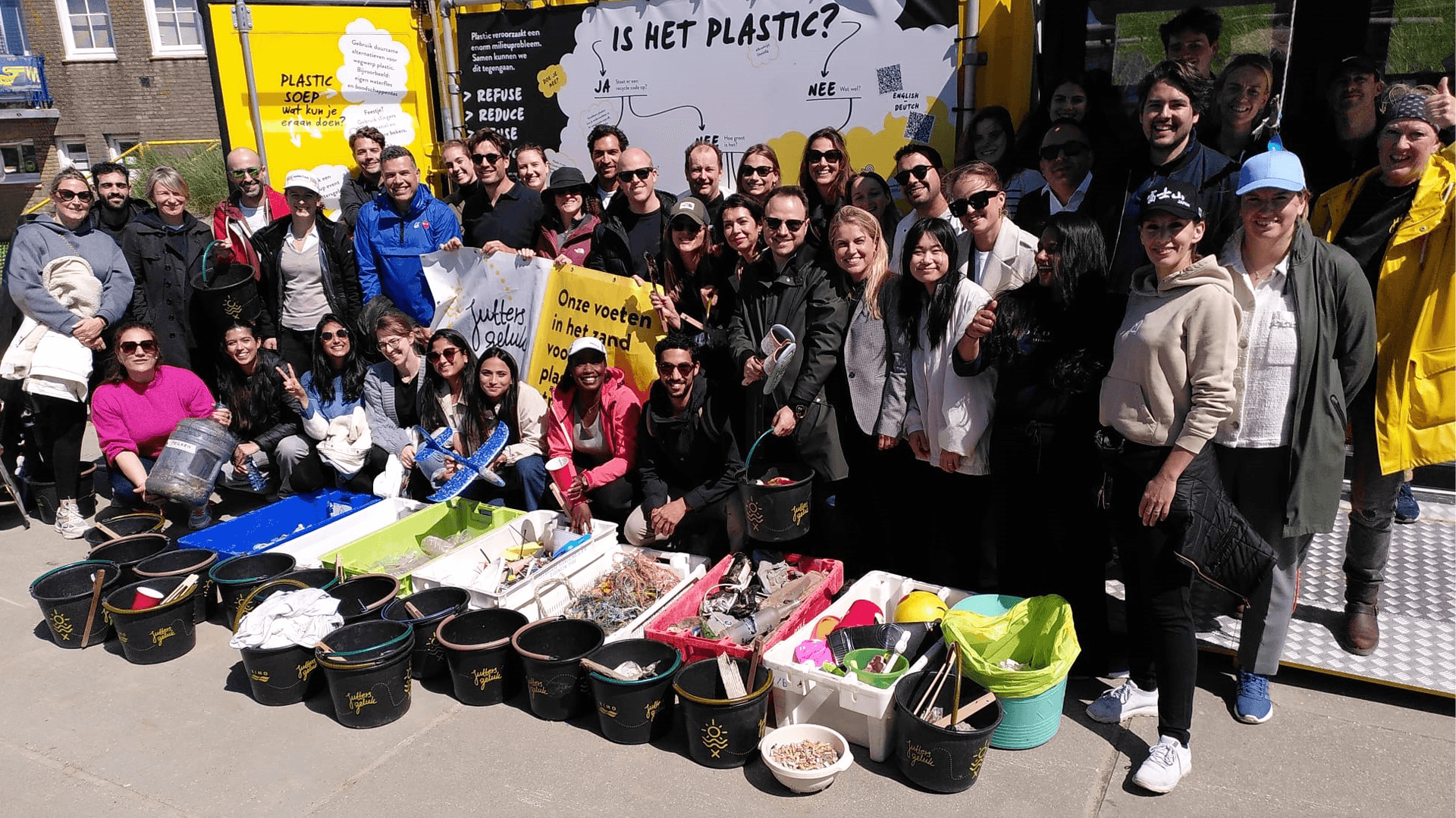 A group of people pose together outdoors in front of a display about plastic. Various plastic items and containers are arranged on the ground in front of them.