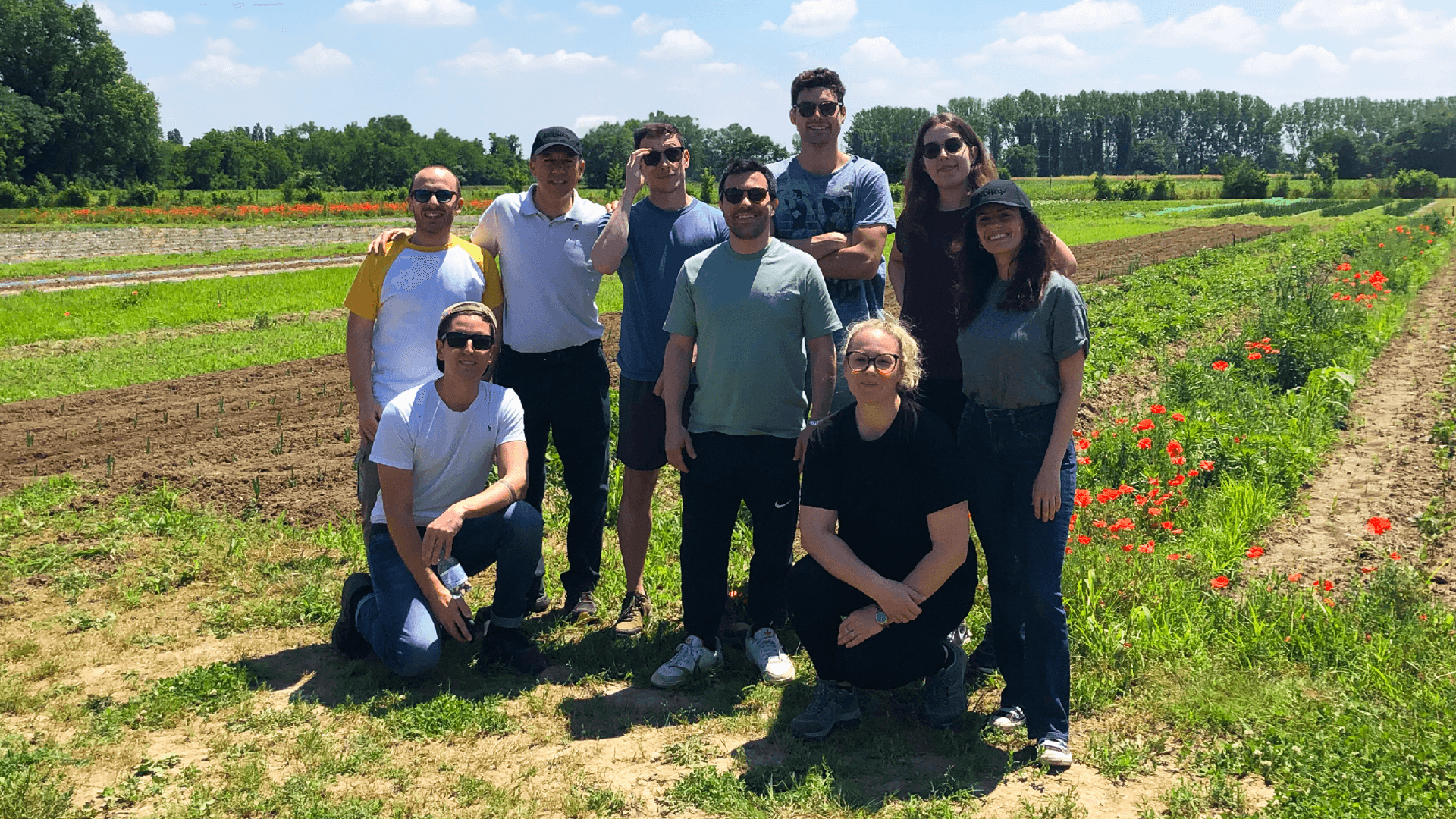 A group of people posing together in a field with rows of crops and flowers on a sunny day.