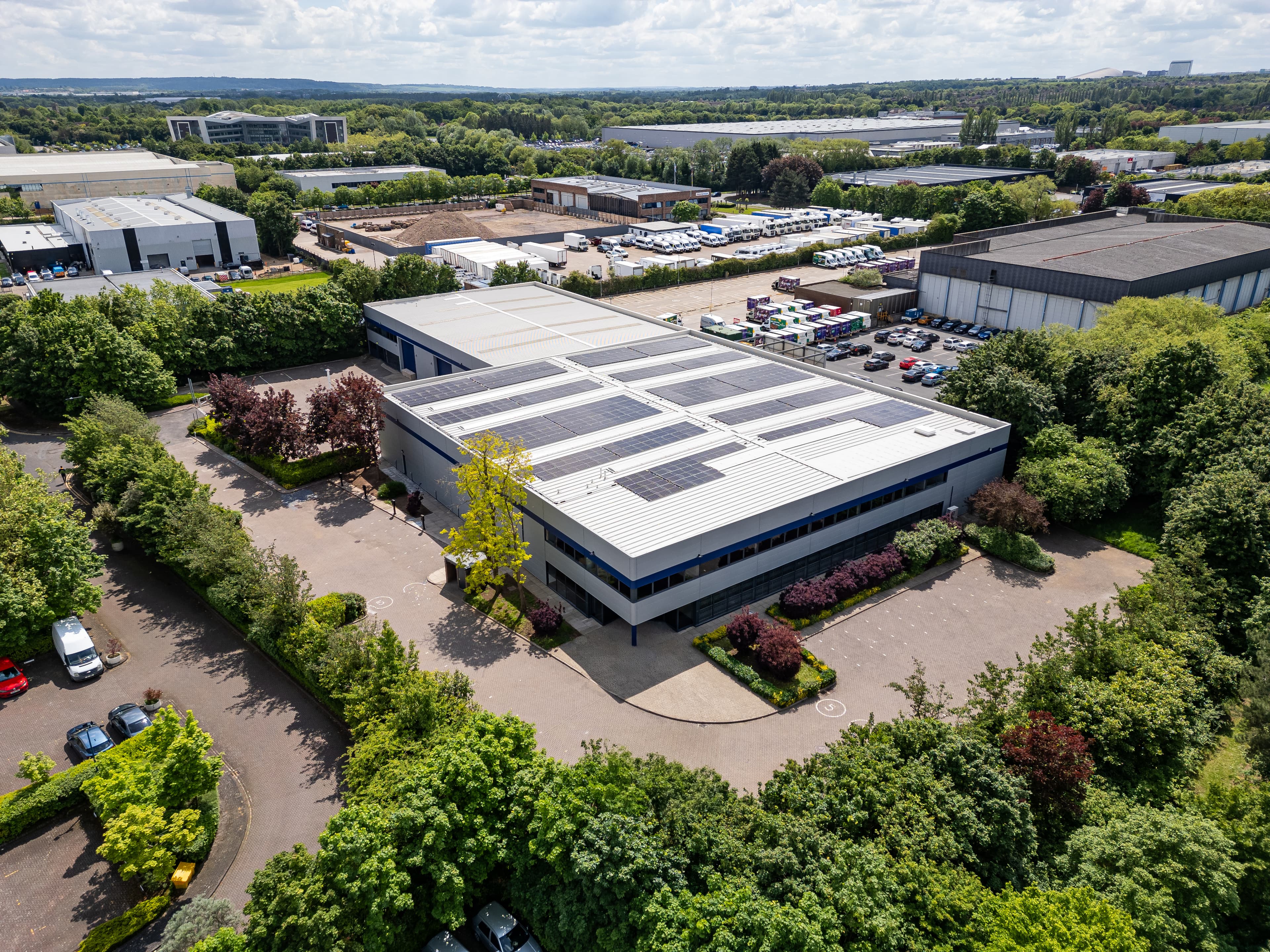 Aerial view of a large warehouse building with solar panels on the roof, surrounded by trees and parking lots. Industrial area and other buildings are visible in the background.
