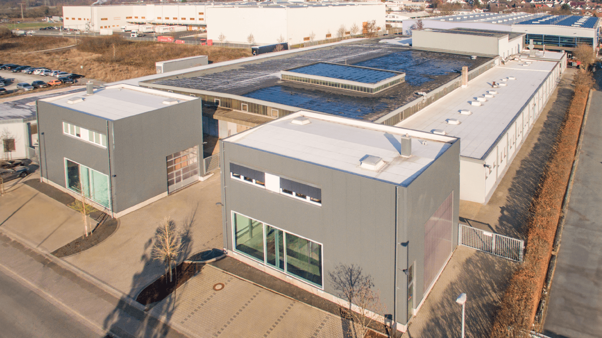 Aerial view of a modern industrial building complex with flat roofs, surrounded by parking areas and roads.