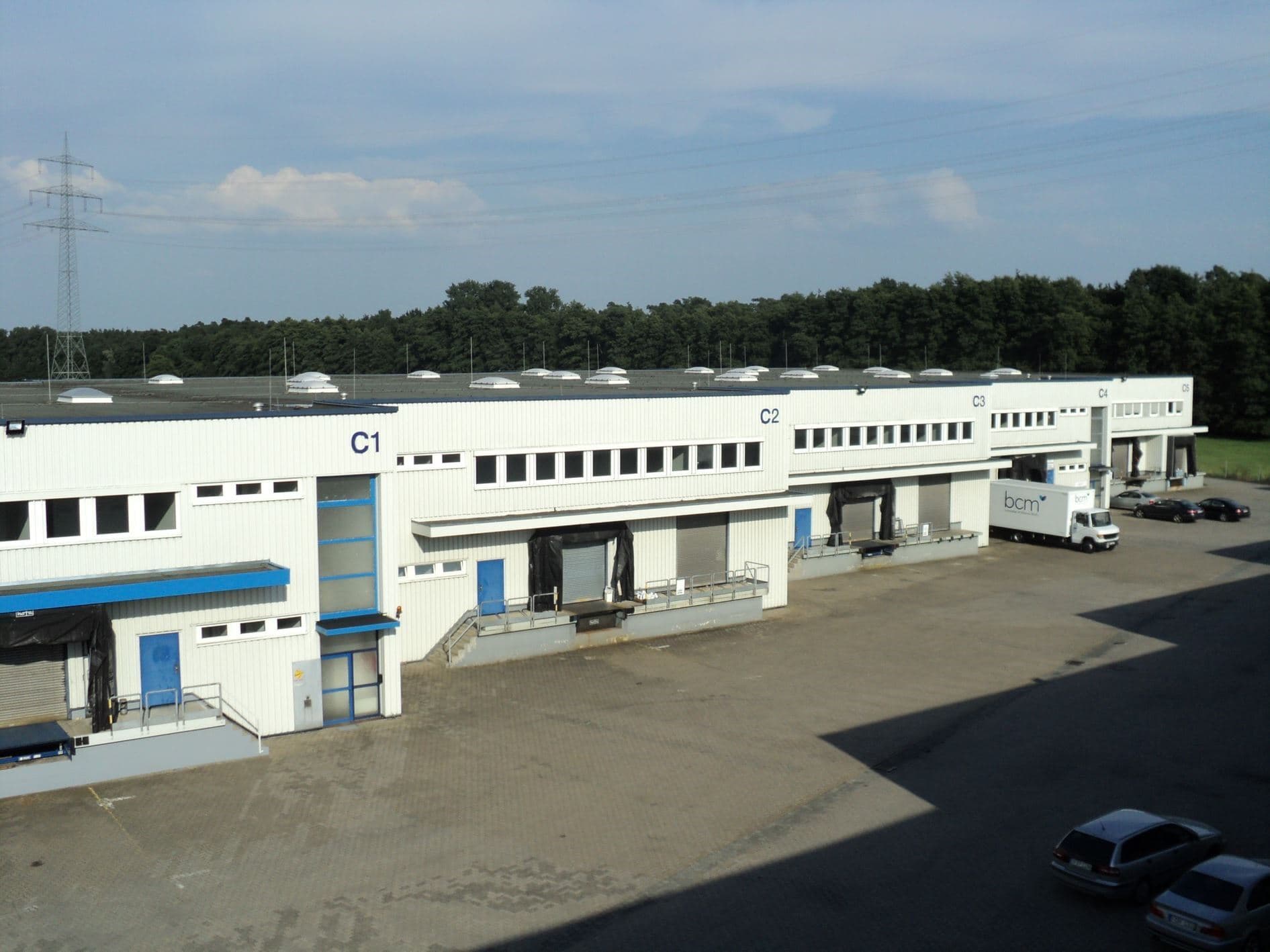 A row of warehouse loading docks with labeled doors C1, C2, and C3. The area is paved, and there are a few parked cars. Trees and a power line tower are visible in the background.