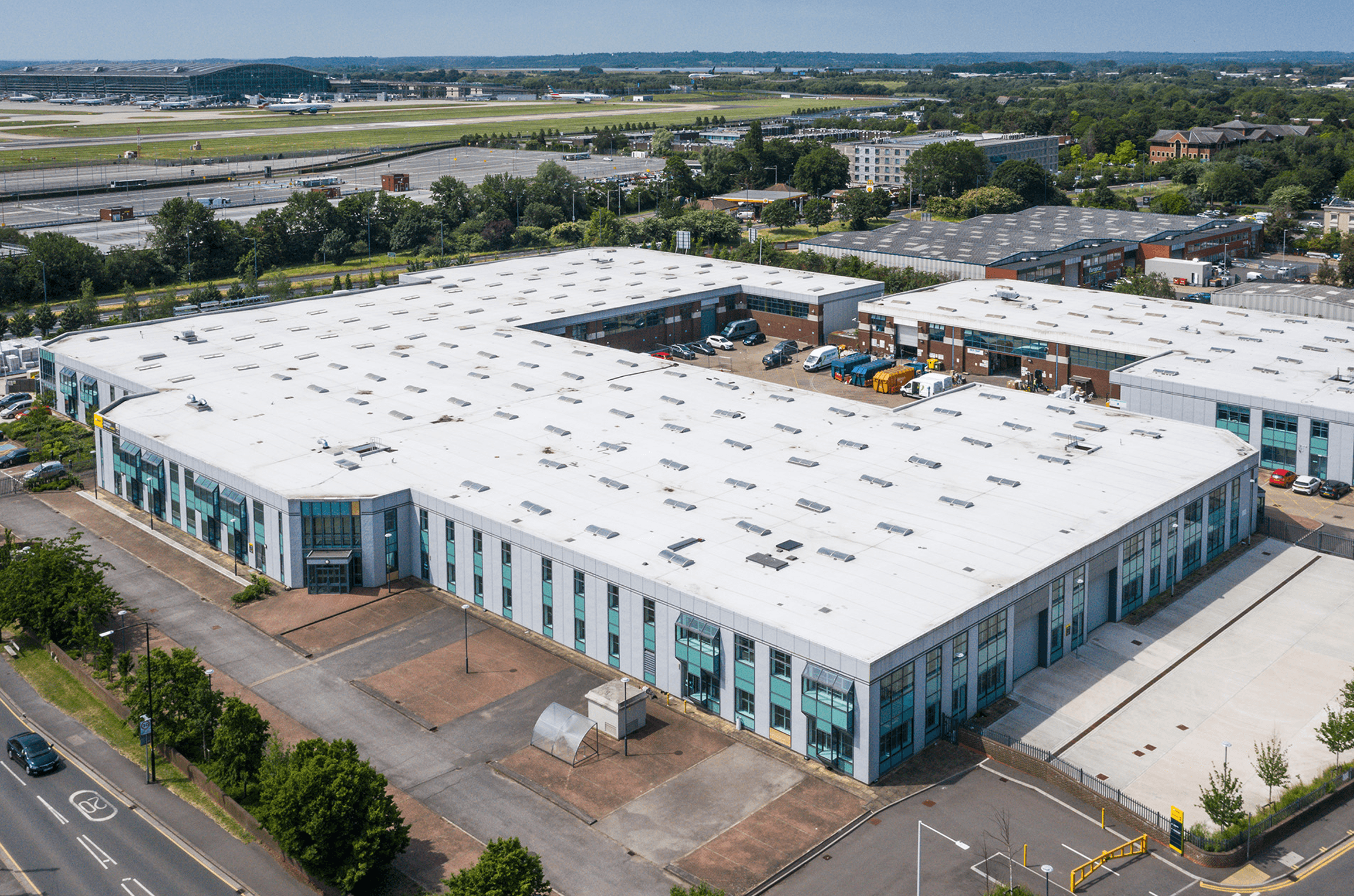 Aerial view of a large industrial complex with white roofs, surrounded by roads and greenery, near an airport runway in the background.