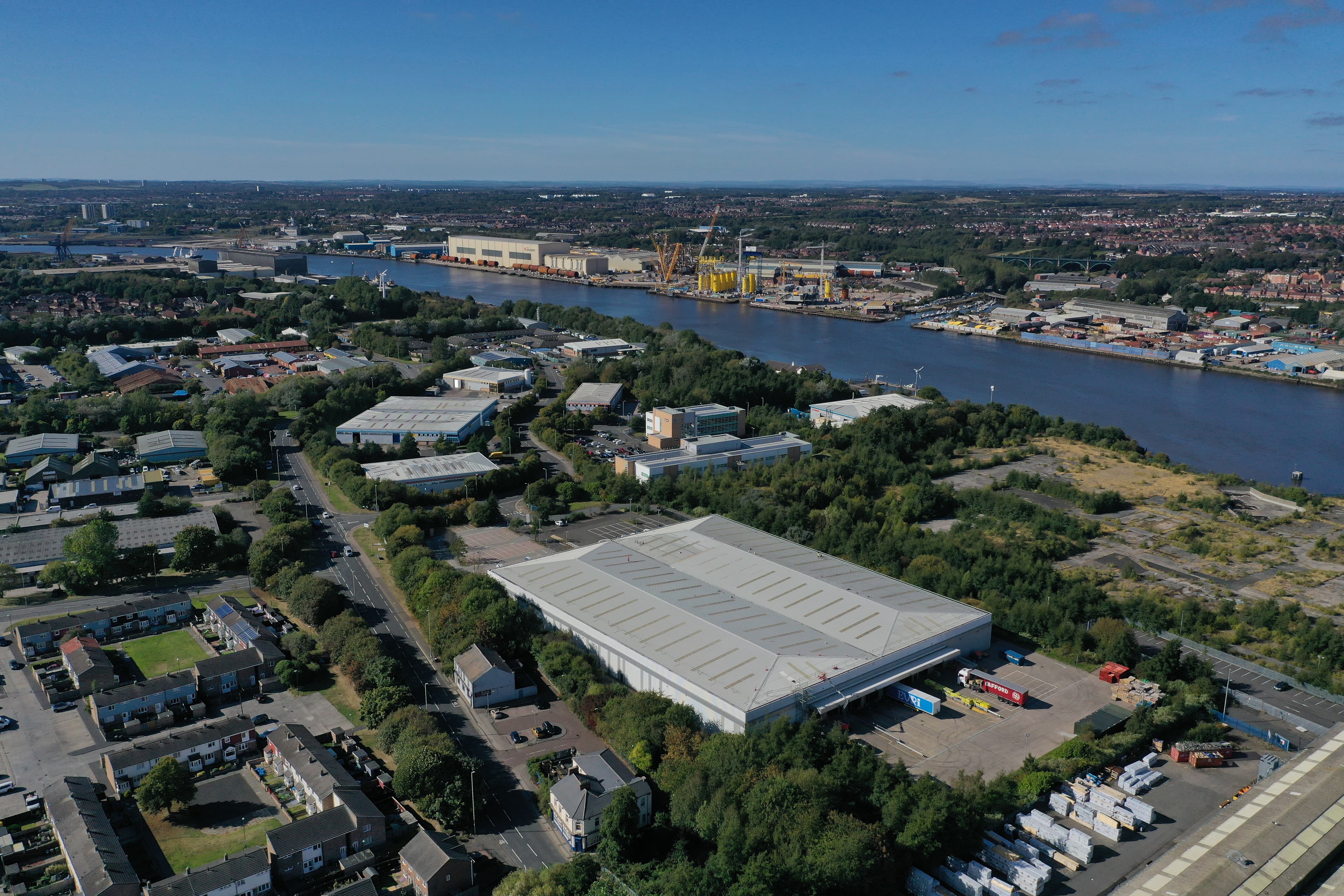 Aerial view of an industrial area beside a river, featuring warehouses, industrial buildings, and cranes. Residential neighborhoods and greenery surround the area, extending into the distance.
