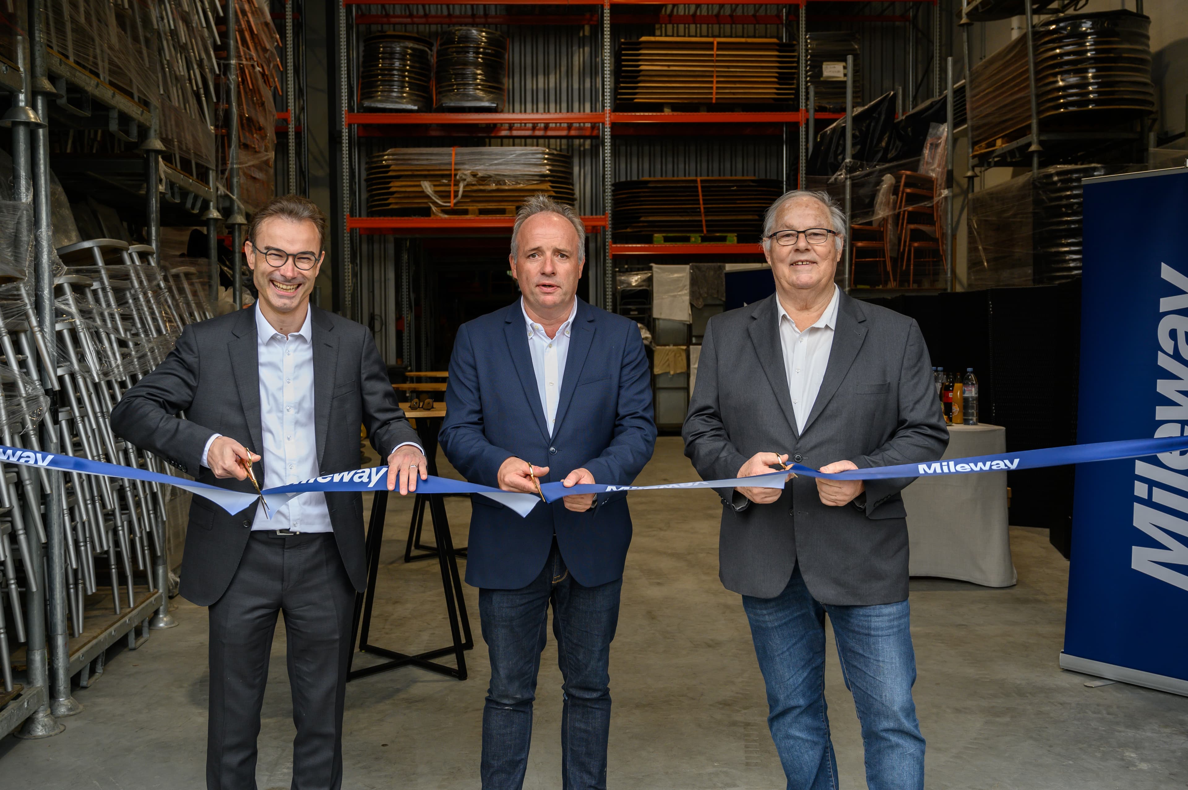 Three men in suits cut a blue ribbon at an indoor event, with stacks of chairs and shelves in the background.