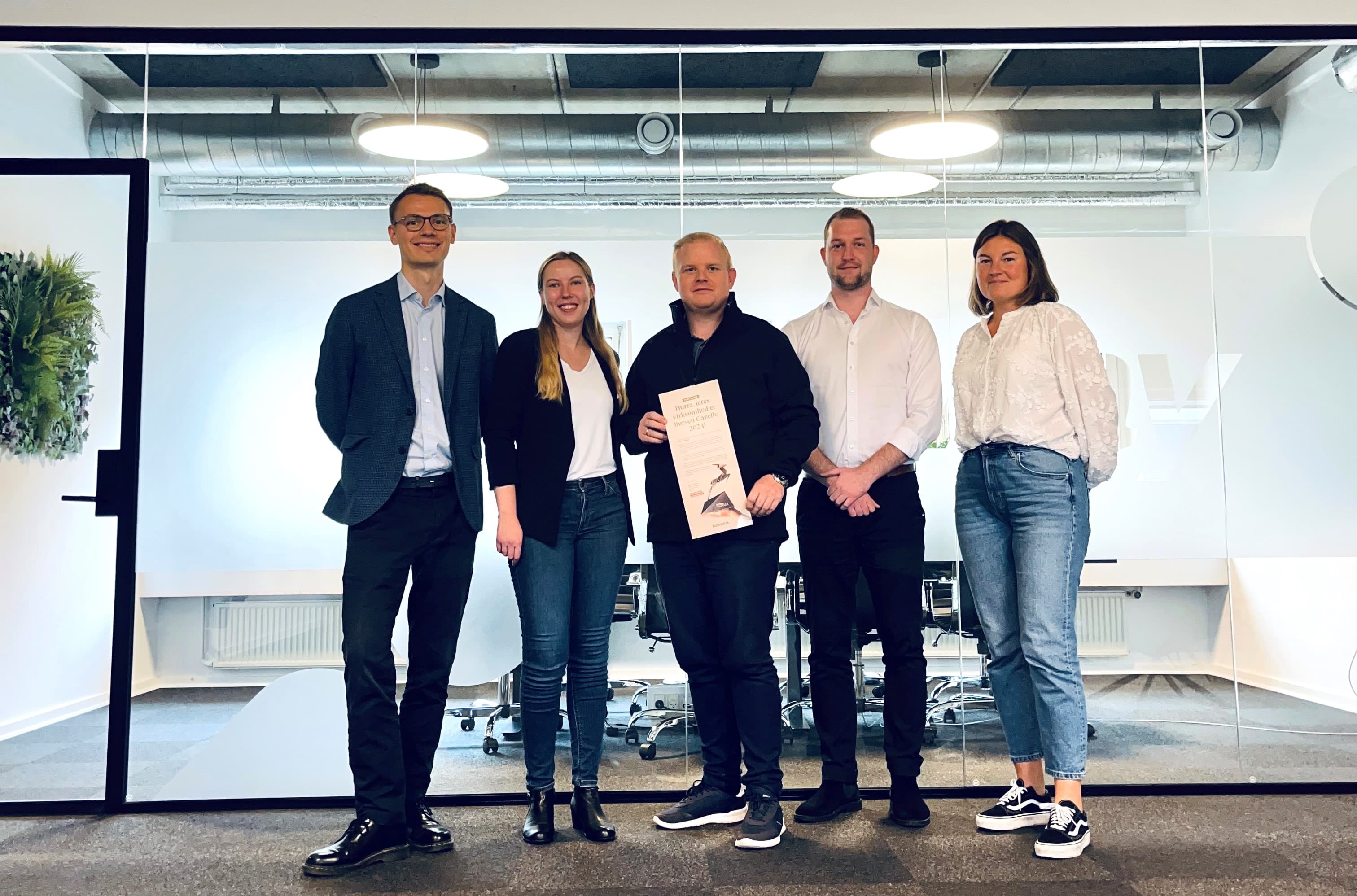 Five people standing in an office, with one holding a framed document. They are posed in front of a glass-walled meeting room.
